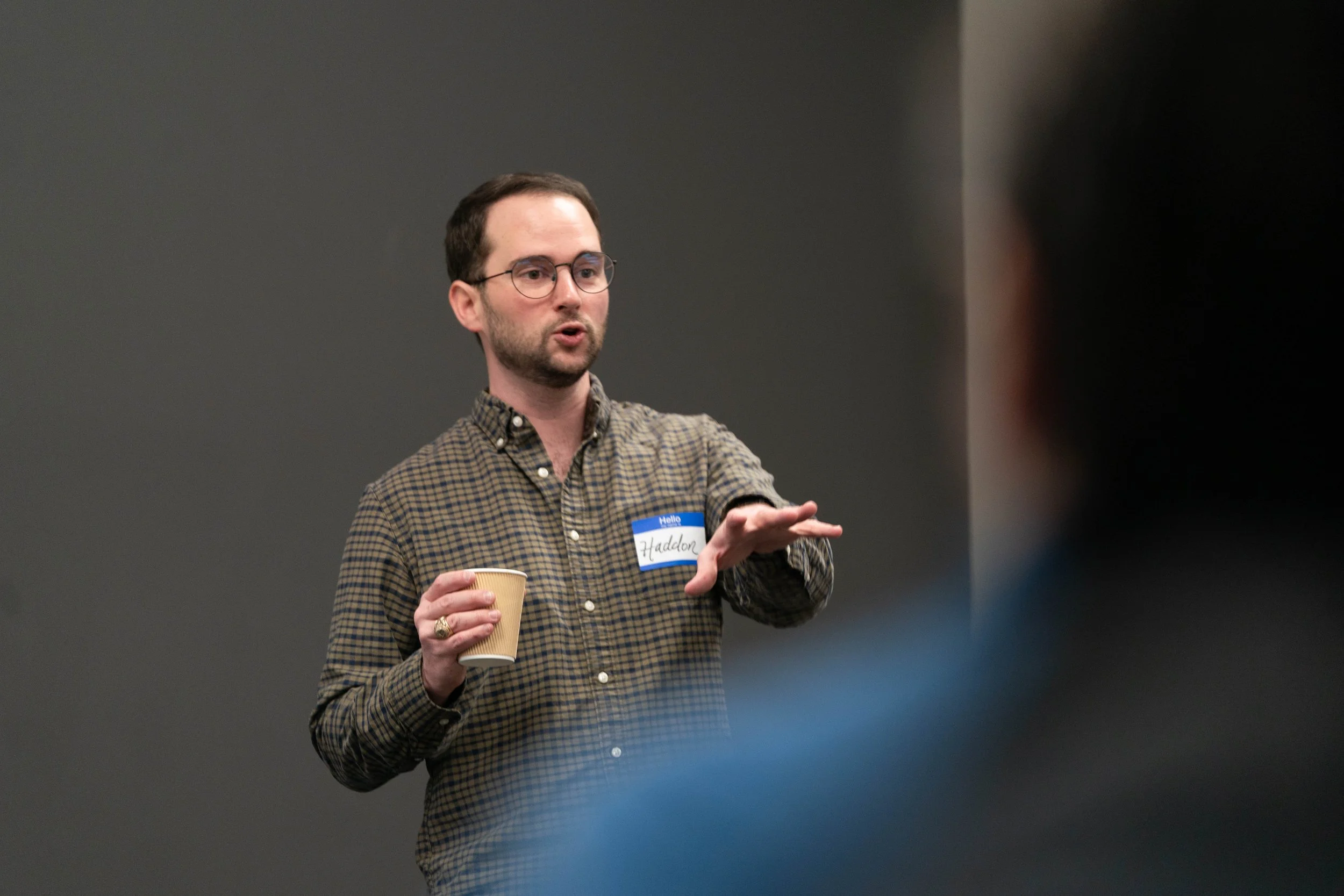 Man with glasses and a beard wearing a plaid shirt, holding a paper cup, speaking in front of a dark background.