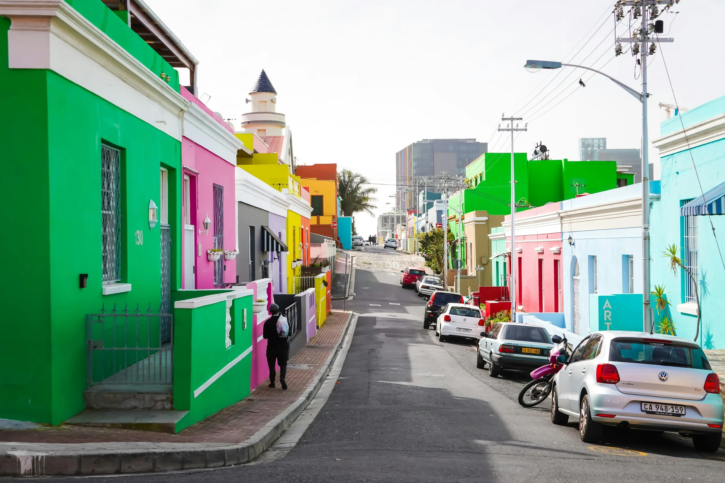 Colorful houses lining a street with cars parked along the curb, including a person walking on the sidewalk.