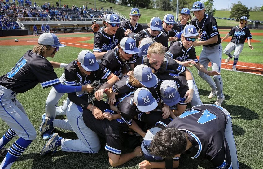 Youth baseball team celebrating a victory in a pile on the field, wearing black and blue uniforms and caps with an 'M' logo.