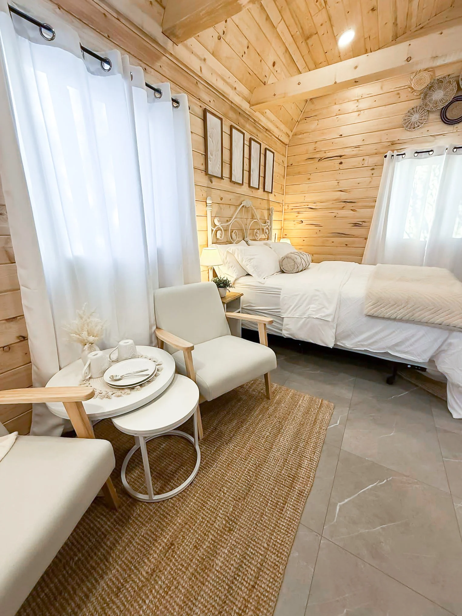 Cozy bedroom with wood-paneled walls and ceiling, white curtains, a white bed, and a small sitting area with a table, chairs, and decorative items.