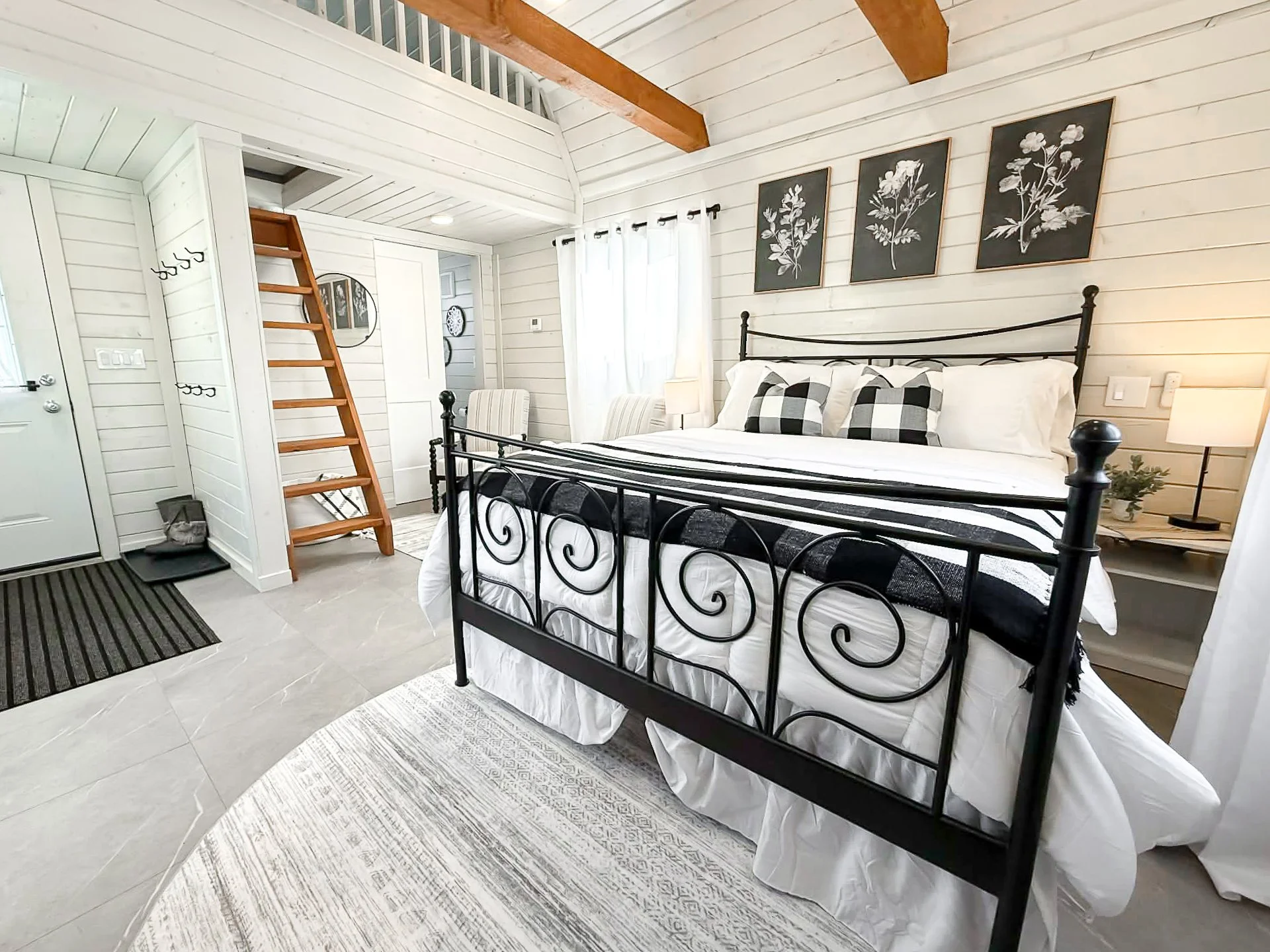Modern bedroom with white wooden walls and ceiling, black metal bed frame with white bedding and black-and-white pillows, three black botanical art pieces on the wall, wooden ladder leading to a loft, nightstand with a lamp and small plant, striped rug on the floor, and a window with curtains.