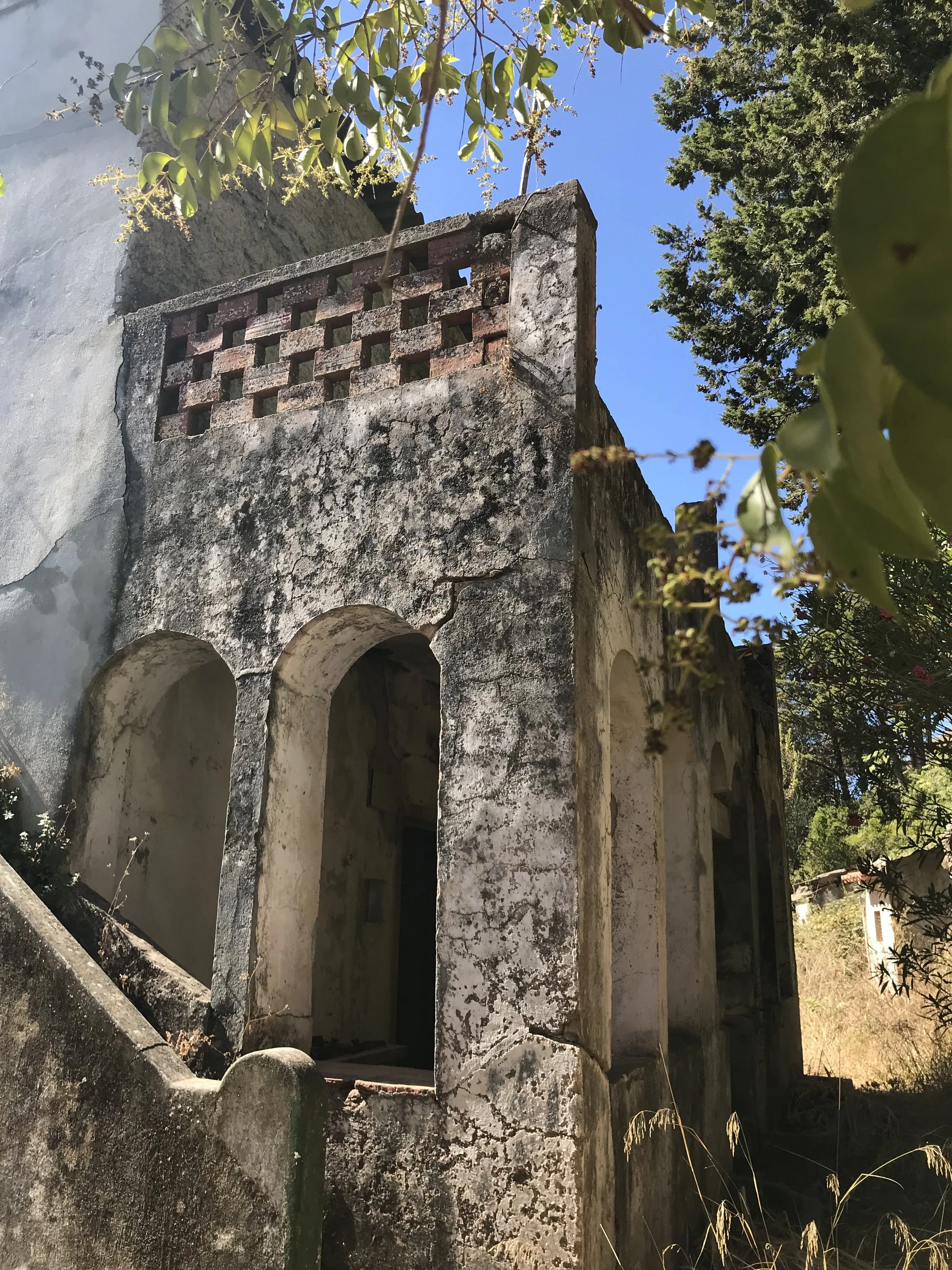 The old quinta da misericordia building, weathered building with arched doorways and a decorative brick lattice on the upper part, surrounded by trees and plants under a clear blue sky.