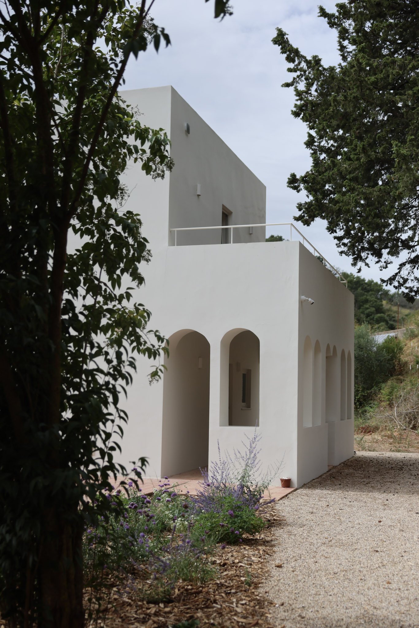 White modern house with arched windows and a terrace, surrounded by trees and plants on a gravel pathway.
