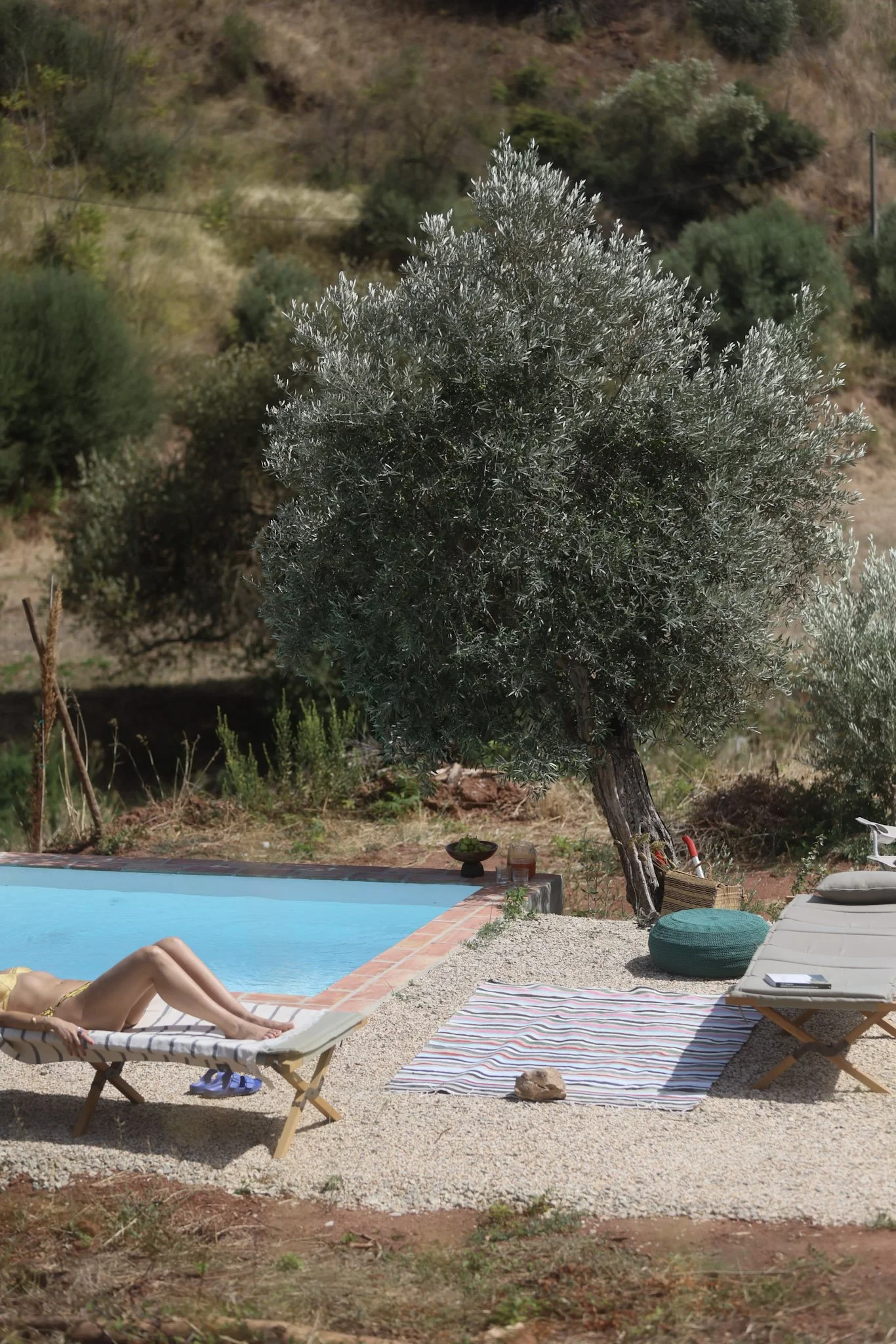A person relaxing on a lounge chair beside a swimming pool in a backyard with a large tree and outdoor furniture.