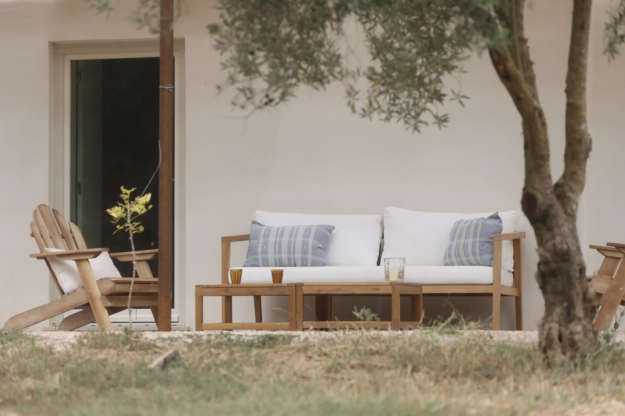 Outdoor patio with a white cushioned wooden sofa with blue and white checkered pillows, surrounded by a tree and wooden chairs, against a white wall.