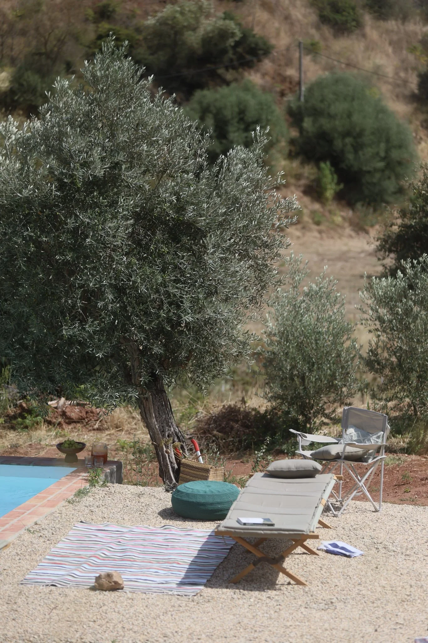 Outdoor patio with a lounge chair, a folding chair, a blanket on the gravel, a small table with drinks, a green pouf, and trees in a dry, hilly landscape.