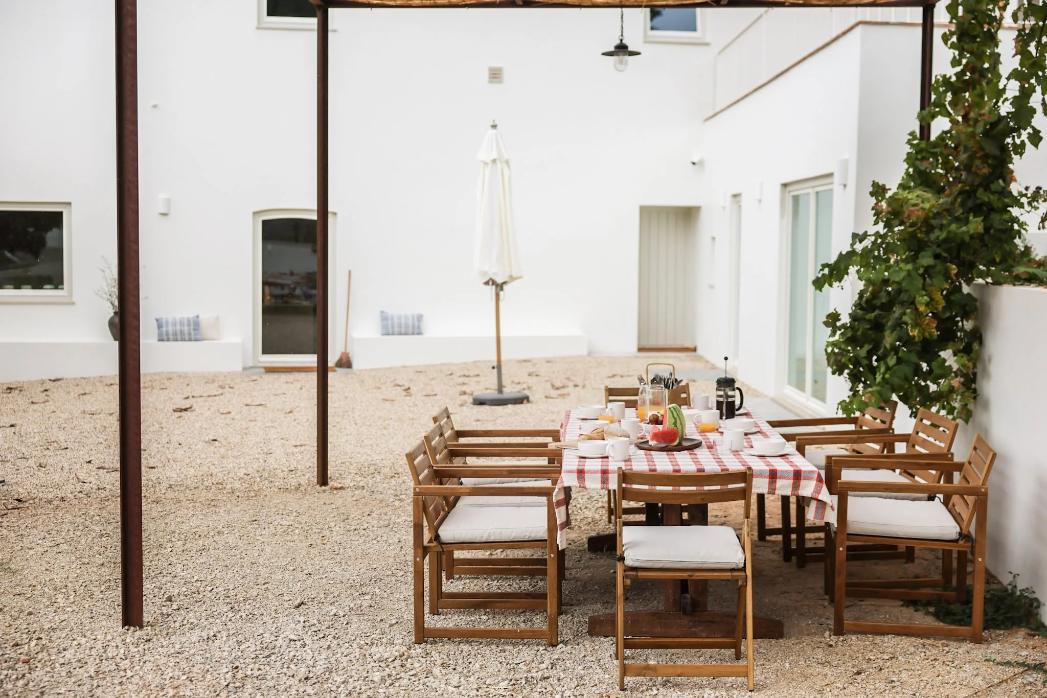 Outdoor patio dining area with a table set with breakfast food, drinks, and watermelon slices, surrounded by wooden chairs with cushions, against white walls with windows and a climbing plant.