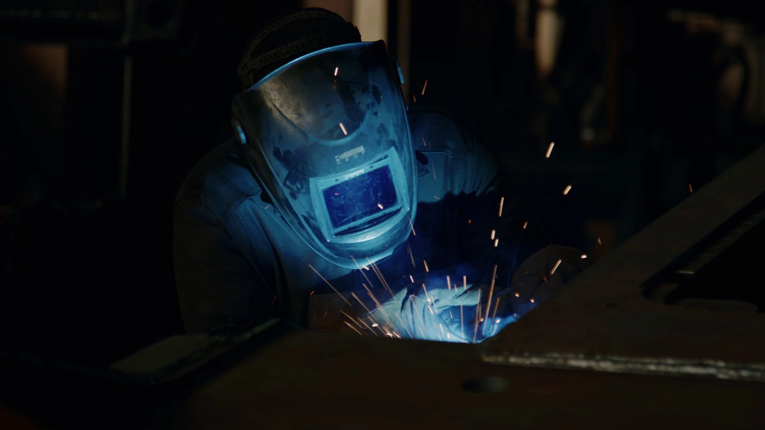 En person i verneutstyr som sveiser metal, med gnister som flyr rundt.