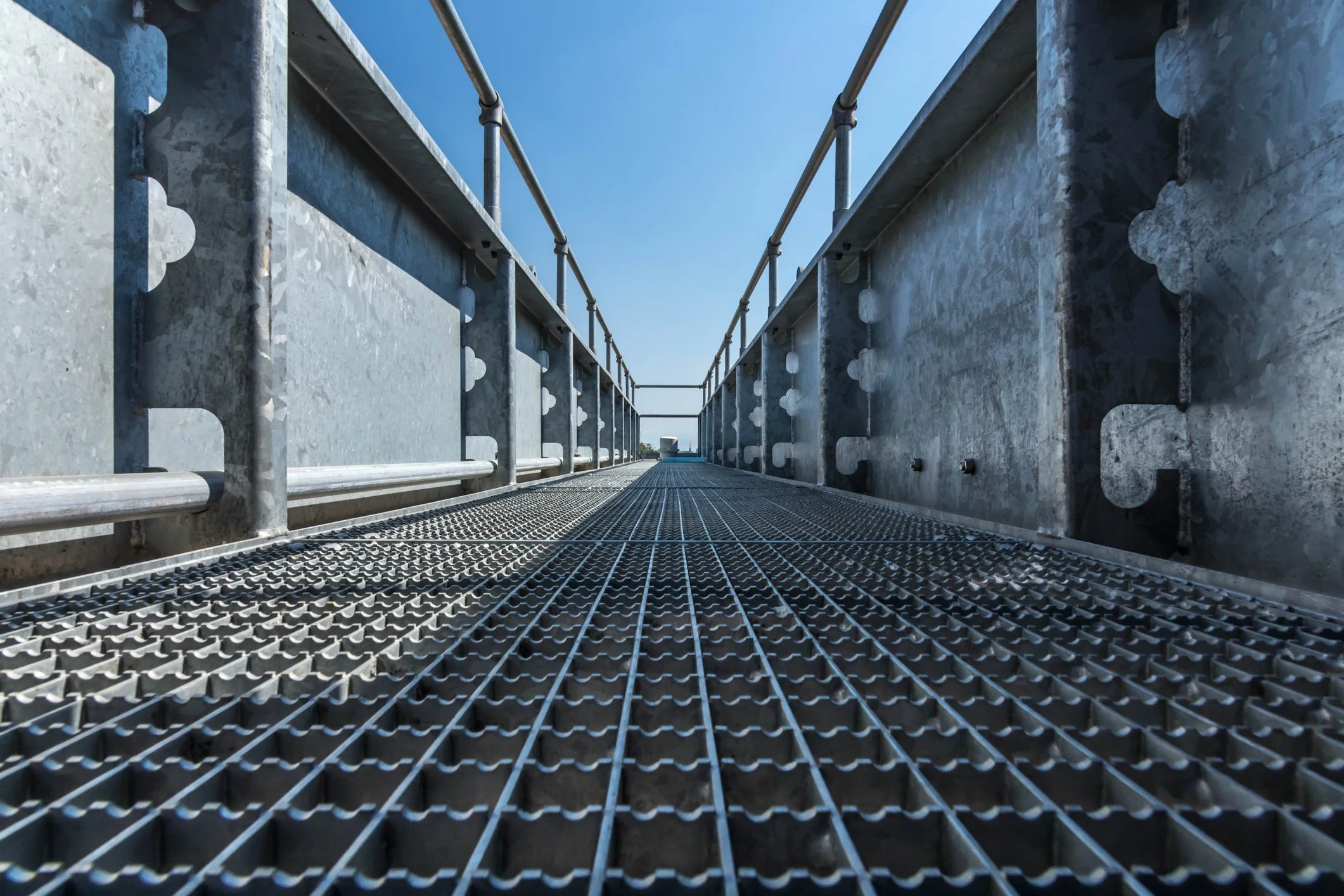 Lavtsynet bilde av en metallbro med sikkerhetsrekkverk og en avløpsrist på gulvet, under en klar blå himmel.