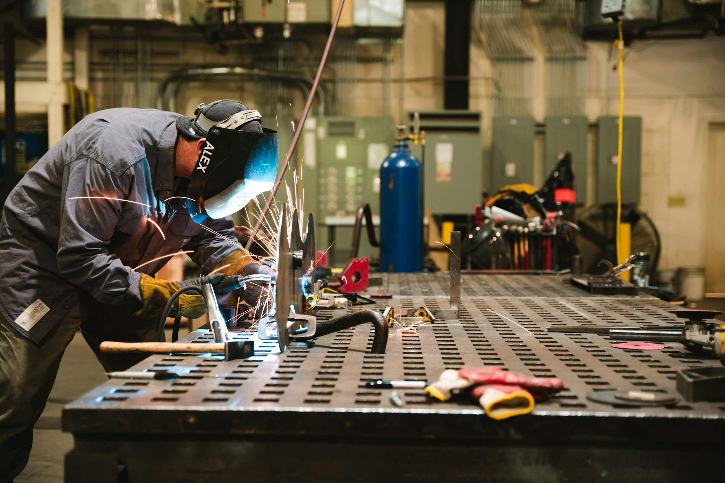 En person som sveiser på et stort metallbord i en industriell fabrikk. Personen bruker verneutstyr, inkludert hjelm og hansker, og det er synlige gnister fra sveiseprosessen.