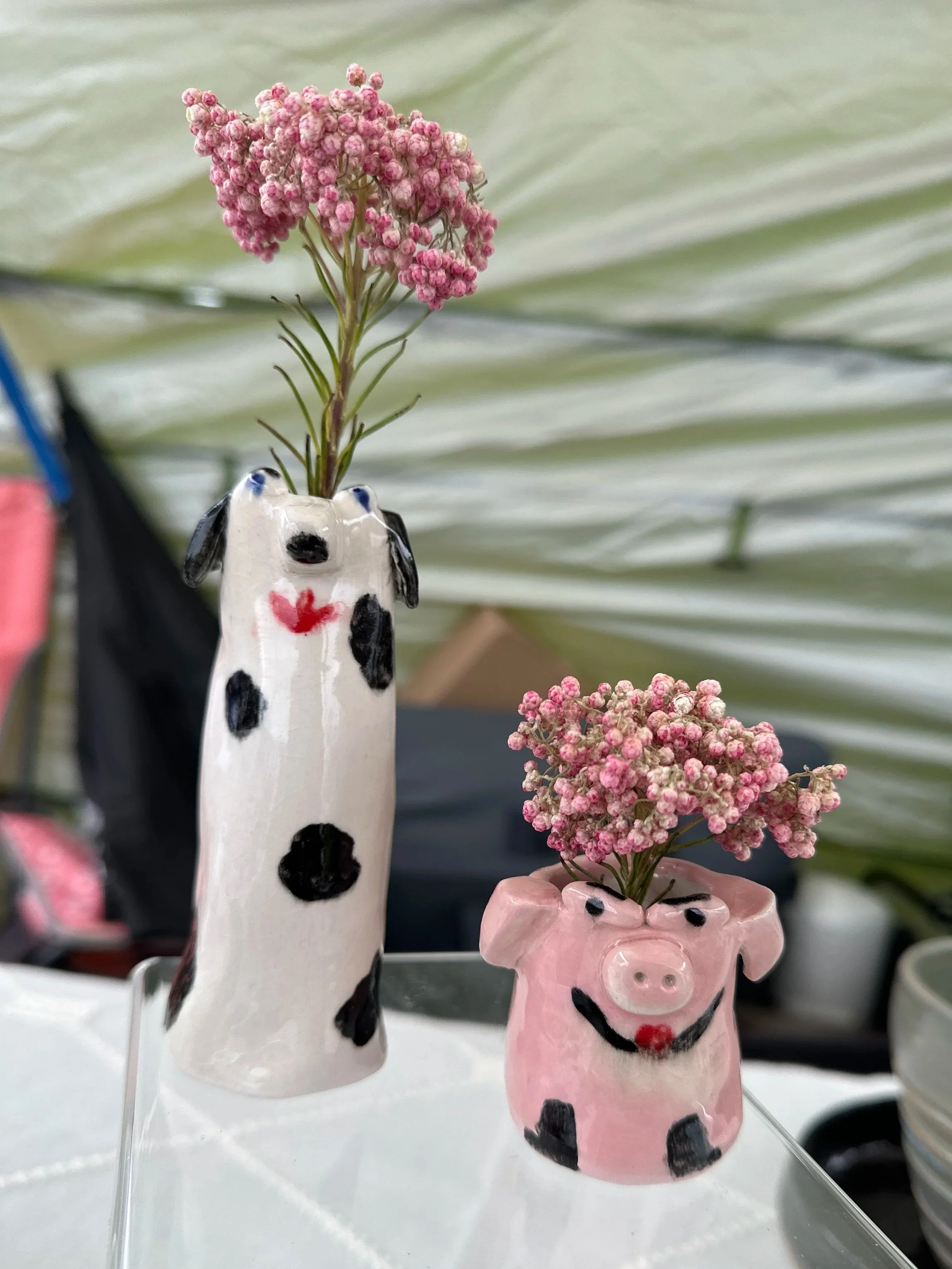 Two animal-shaped ceramic vases, one designed as a cow and the other as a pig, each holding pink flowers, with a striped green and beige background.