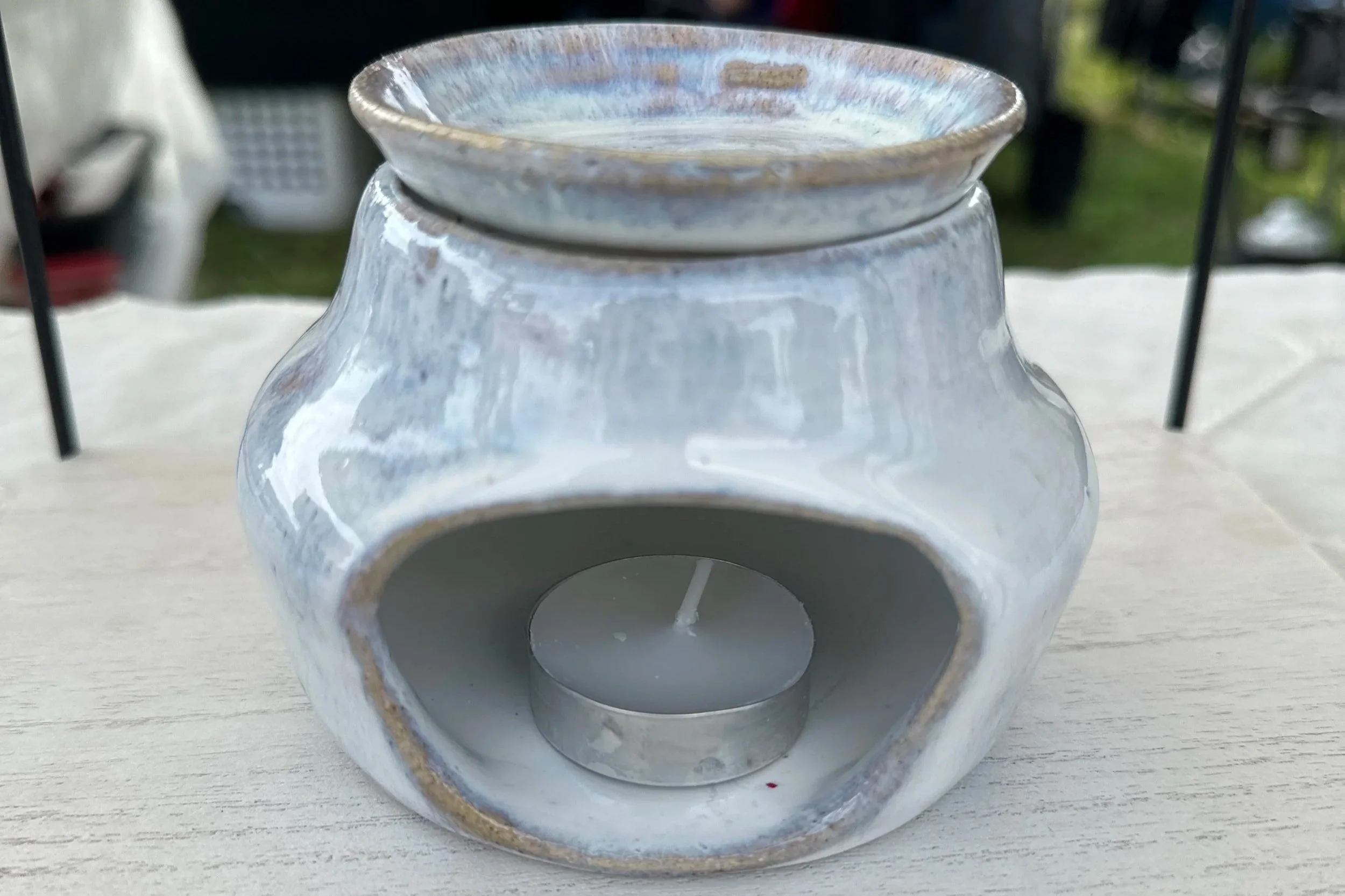A ceramic oil burner with a tealight candle inside, placed on a light-colored wooden surface, outdoors with blurred background.