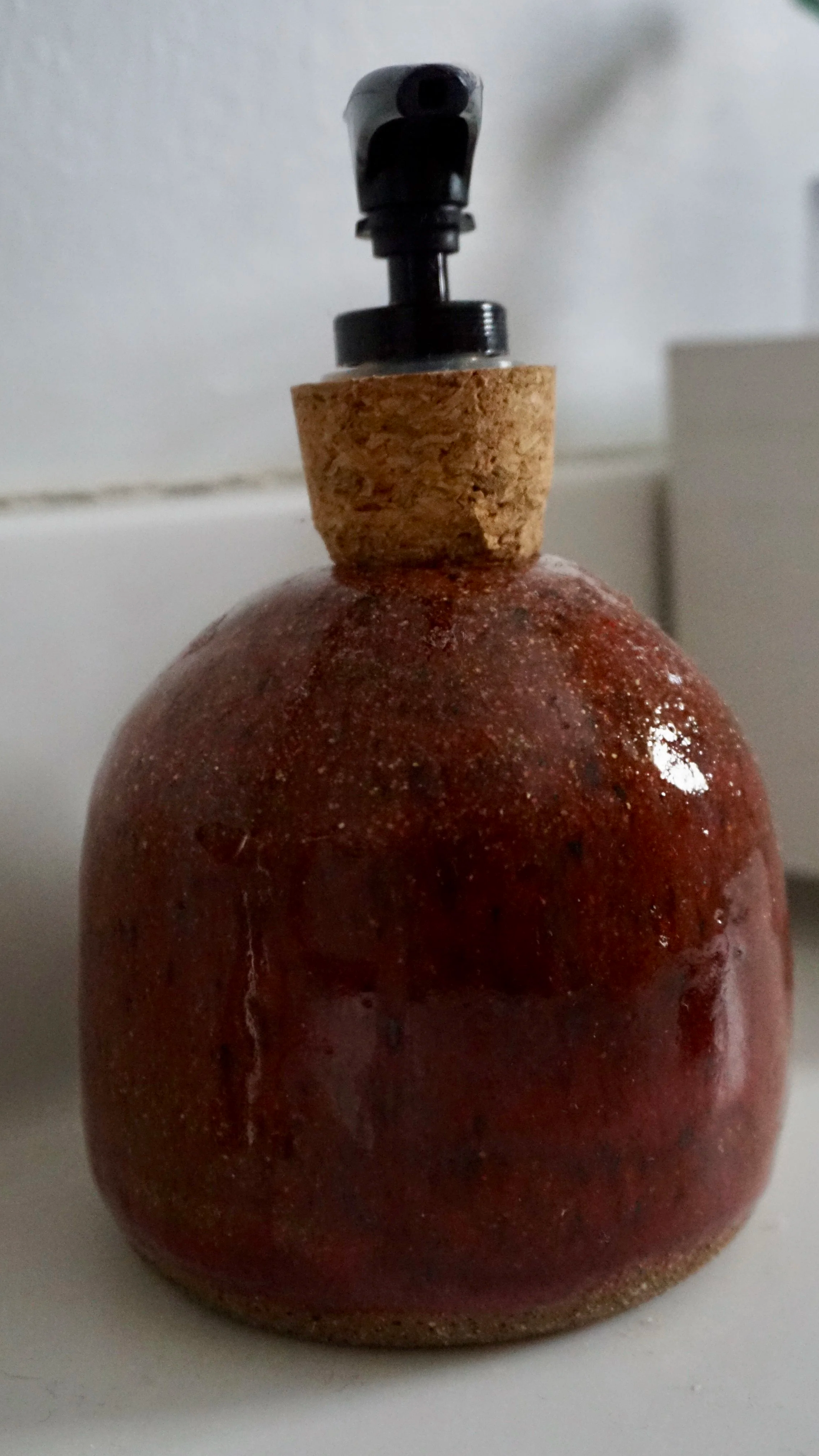 A ceramic decorative bottle with a round, red and brown glazed body, a cork stopper, and a black spray nozzle on top.