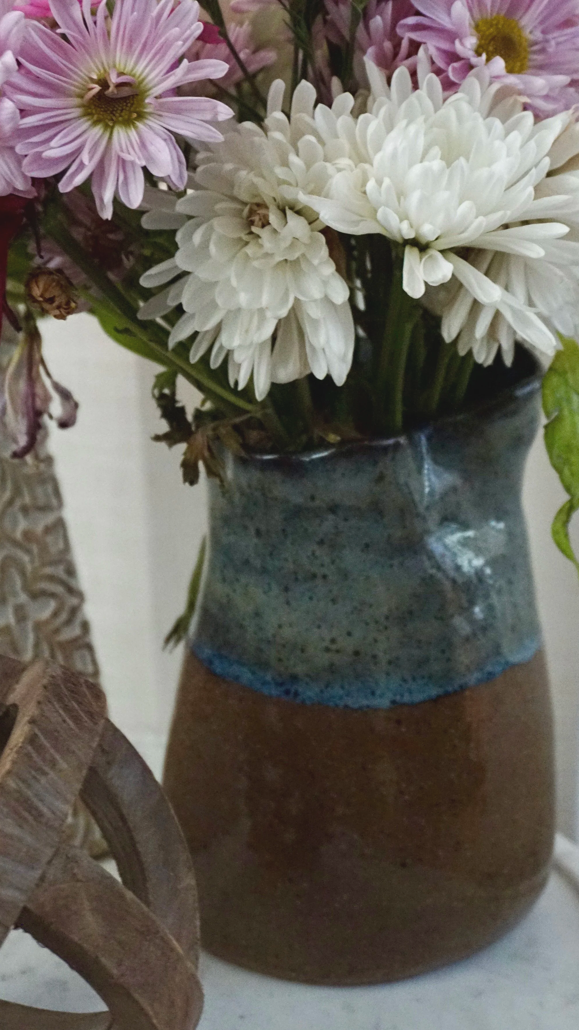 A ceramic vase with white and pink flowers, including chrysanthemums, on a surface next to a dried decorative item.