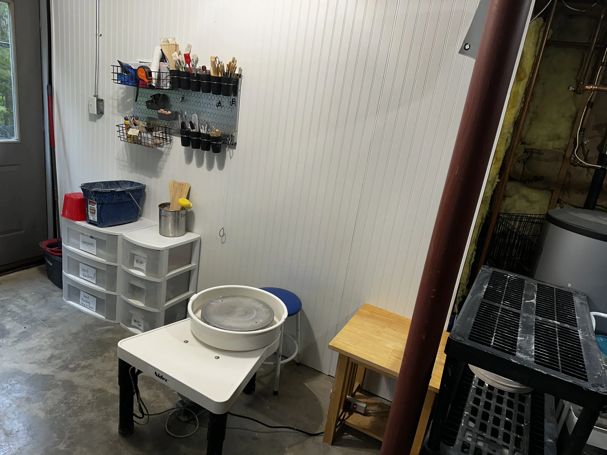 Workshop or craft space with wall-mounted tool organizer, plastic storage drawers labeled for supplies, spinning pottery wheel, and various tools and materials around.