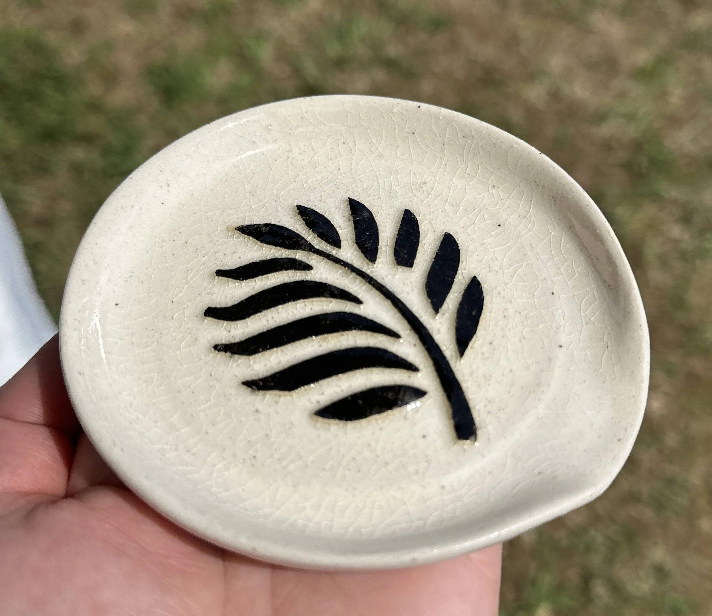 Hand holding a small beige ceramic dish with a black fern leaf design in the center, outdoors on grass.