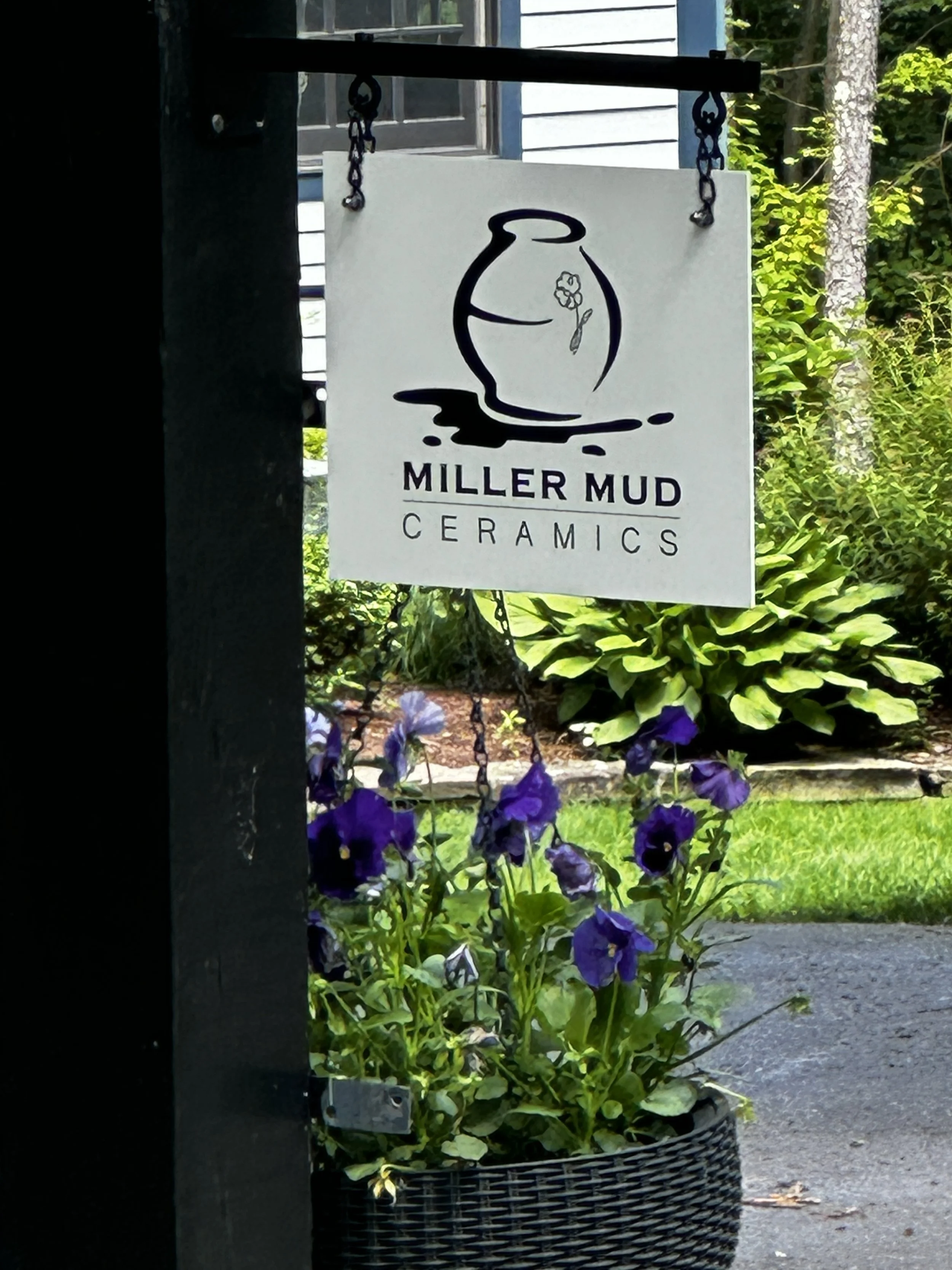 Sign for Miller Mud Ceramics hanging from a black post, with purple flowers in a basket below, outdoor garden with green plants and a house in the background.