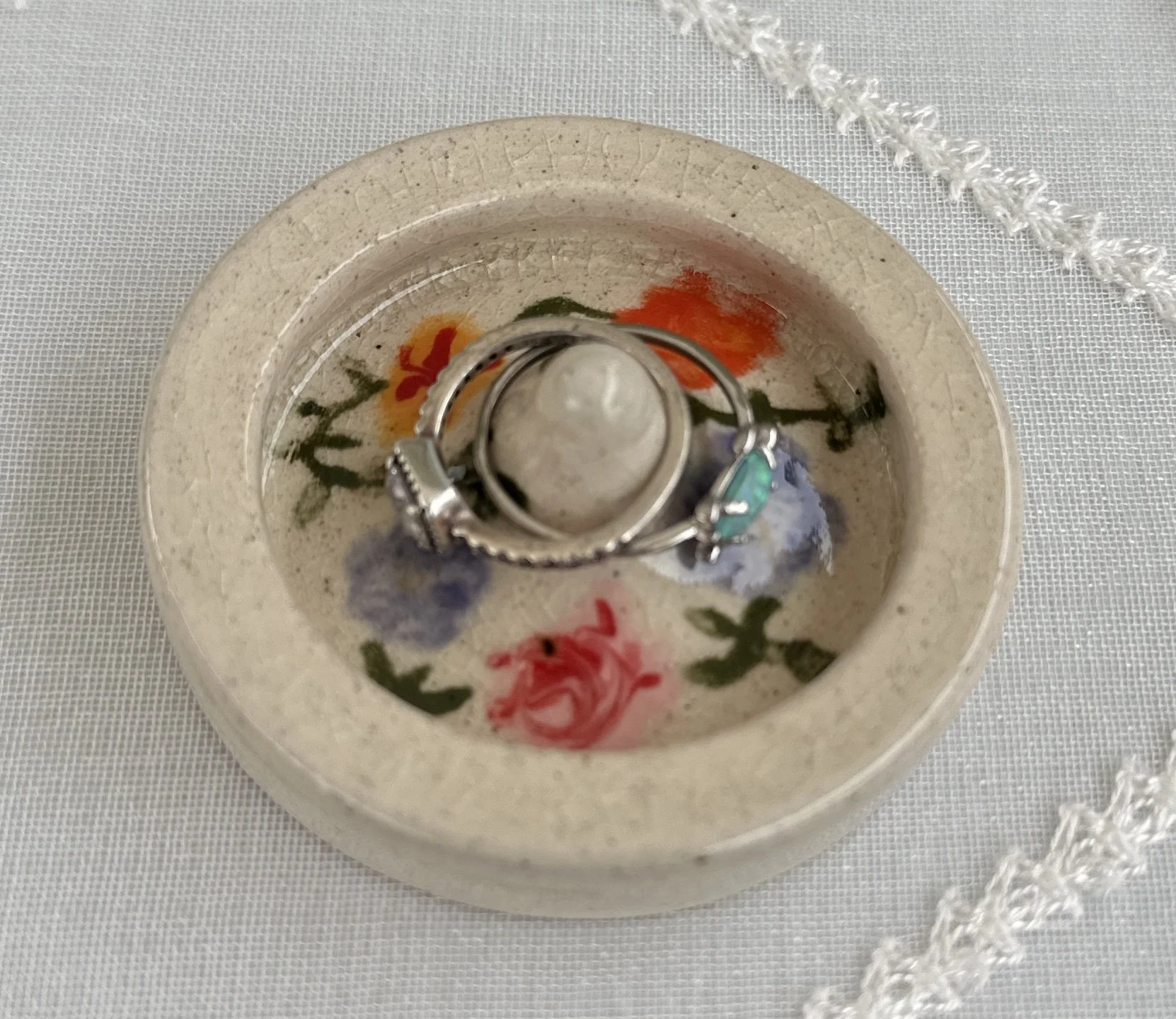 A ring resting on top of other rings inside a small ceramic dish with colorful floral designs, placed on a white fabric surface with a decorative white embroidered trim.