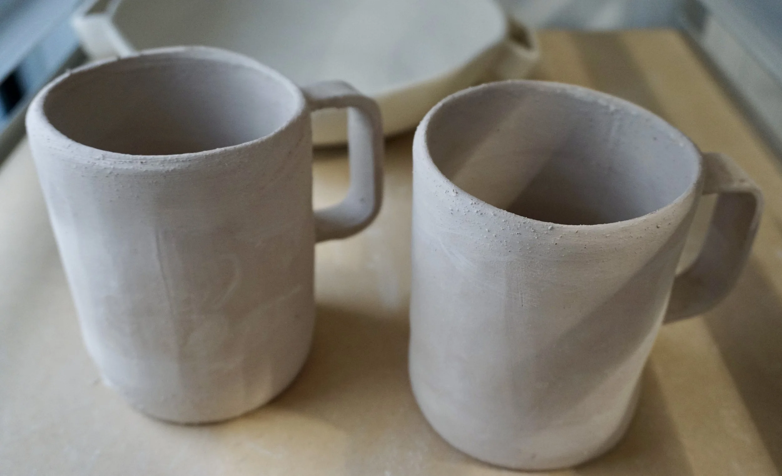 Two unfinished ceramic mugs on a work surface, with one mug in the foreground and the other slightly behind, both with a matte surface and rectangular handles, in a pottery studio.
