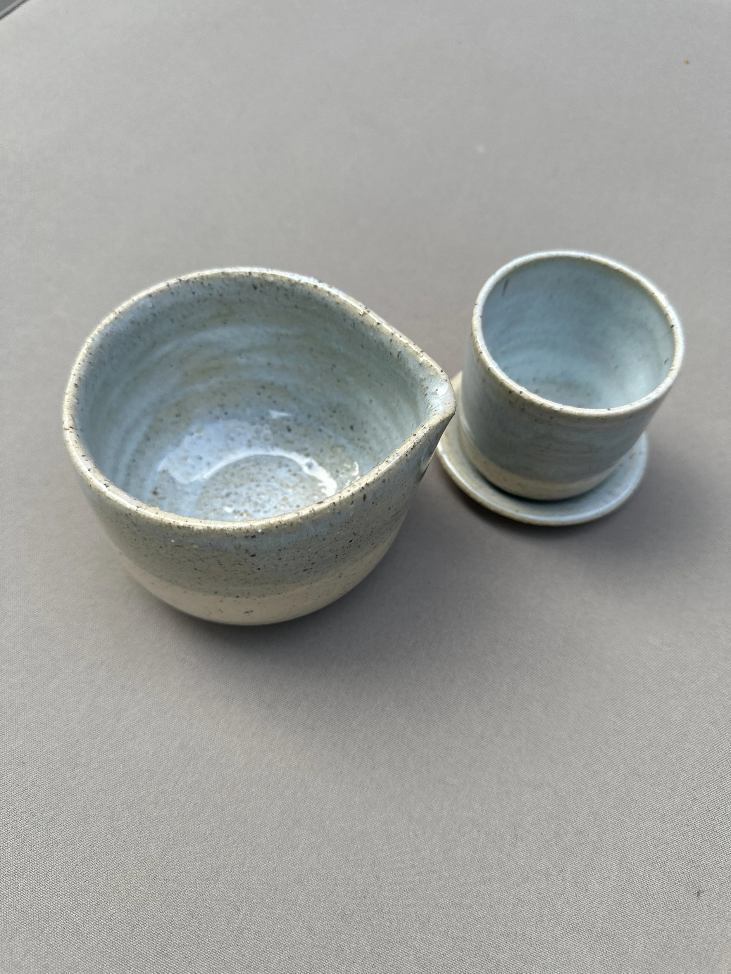 Two small ceramic cups with a light blue glaze, one on a saucer and the other without, placed on a light gray surface.