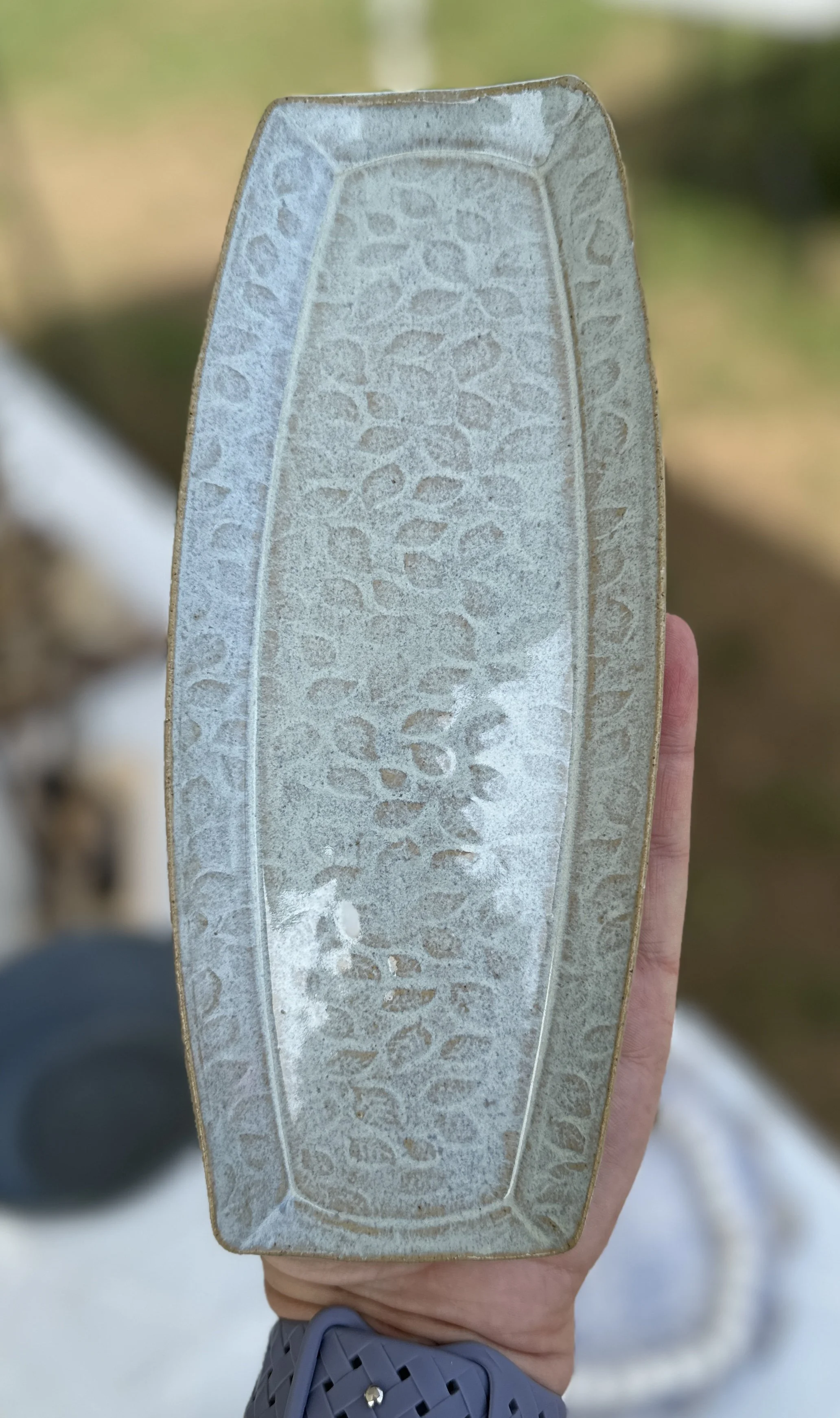 A person's hand holding a rectangular glass with a textured surface, outdoor background with blurred foliage.