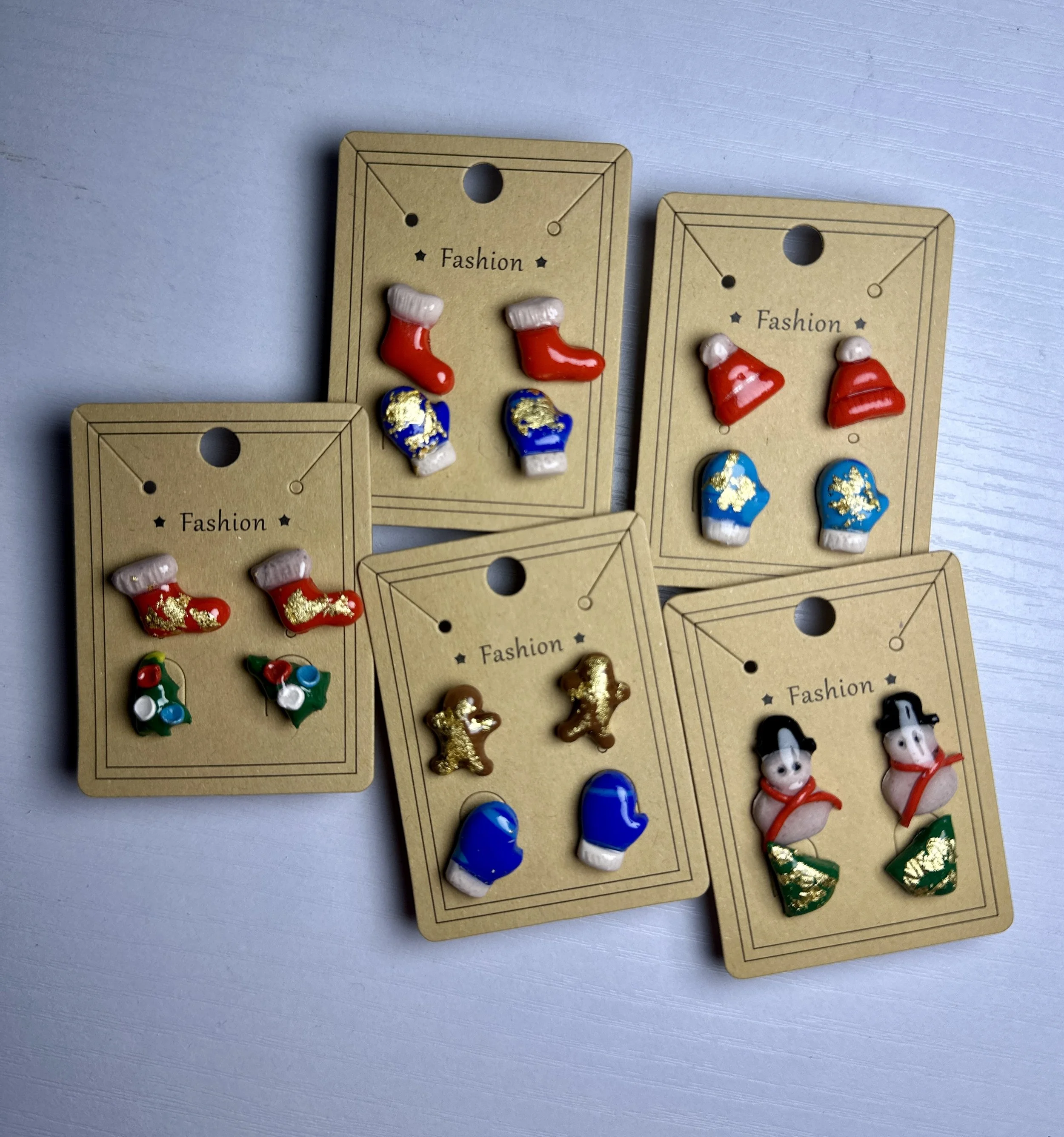 Five pairs of colorful holiday-themed earrings on cardboard cards, each with different festive designs such as stockings, bells, gingerbread men, snowmen, and gloves.