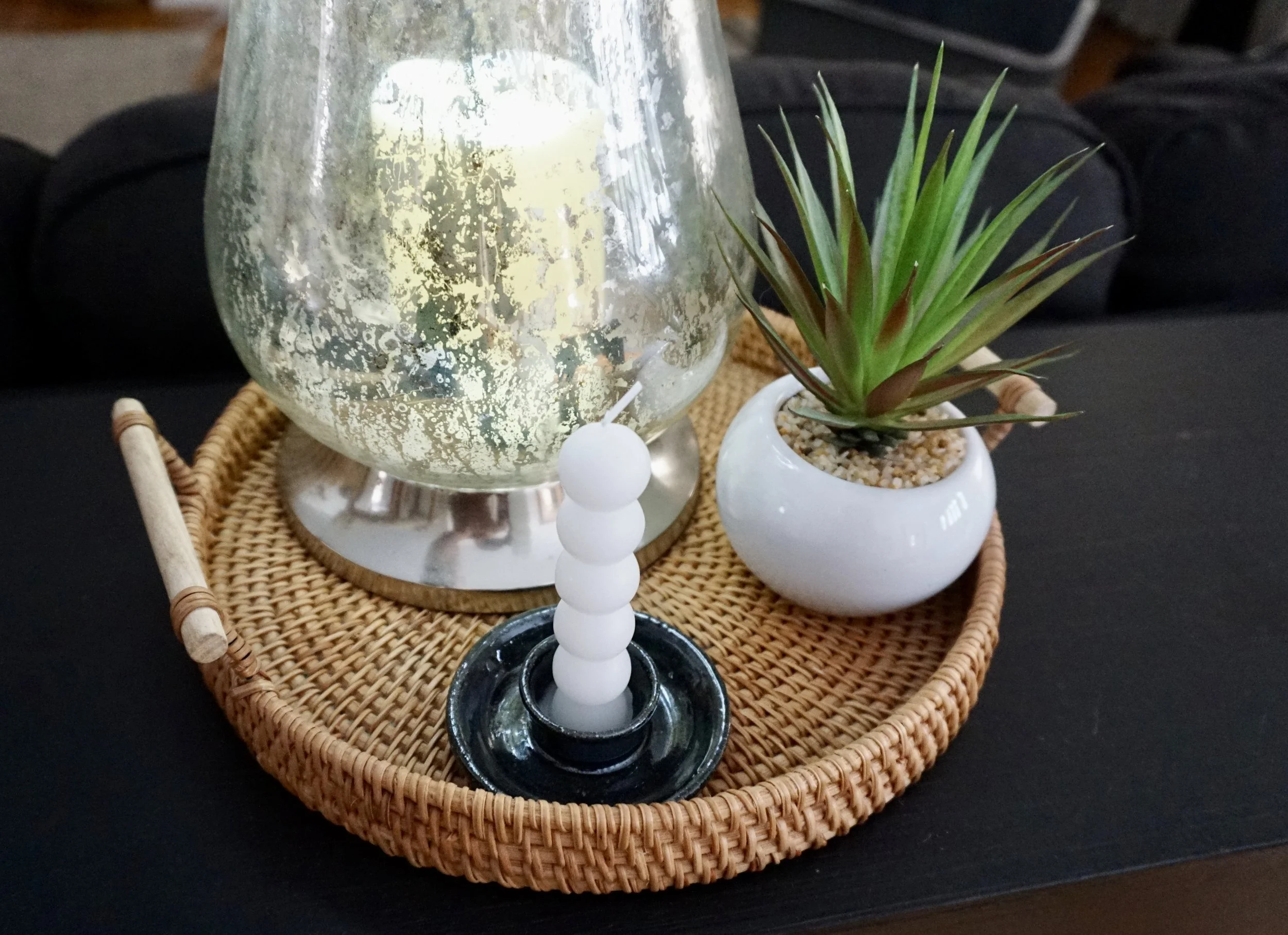 A wicker tray holding a glass candle holder with a white candle, a modern candle snuffer with white balls, and a small white pot with a green succulent plant, all on a black surface.