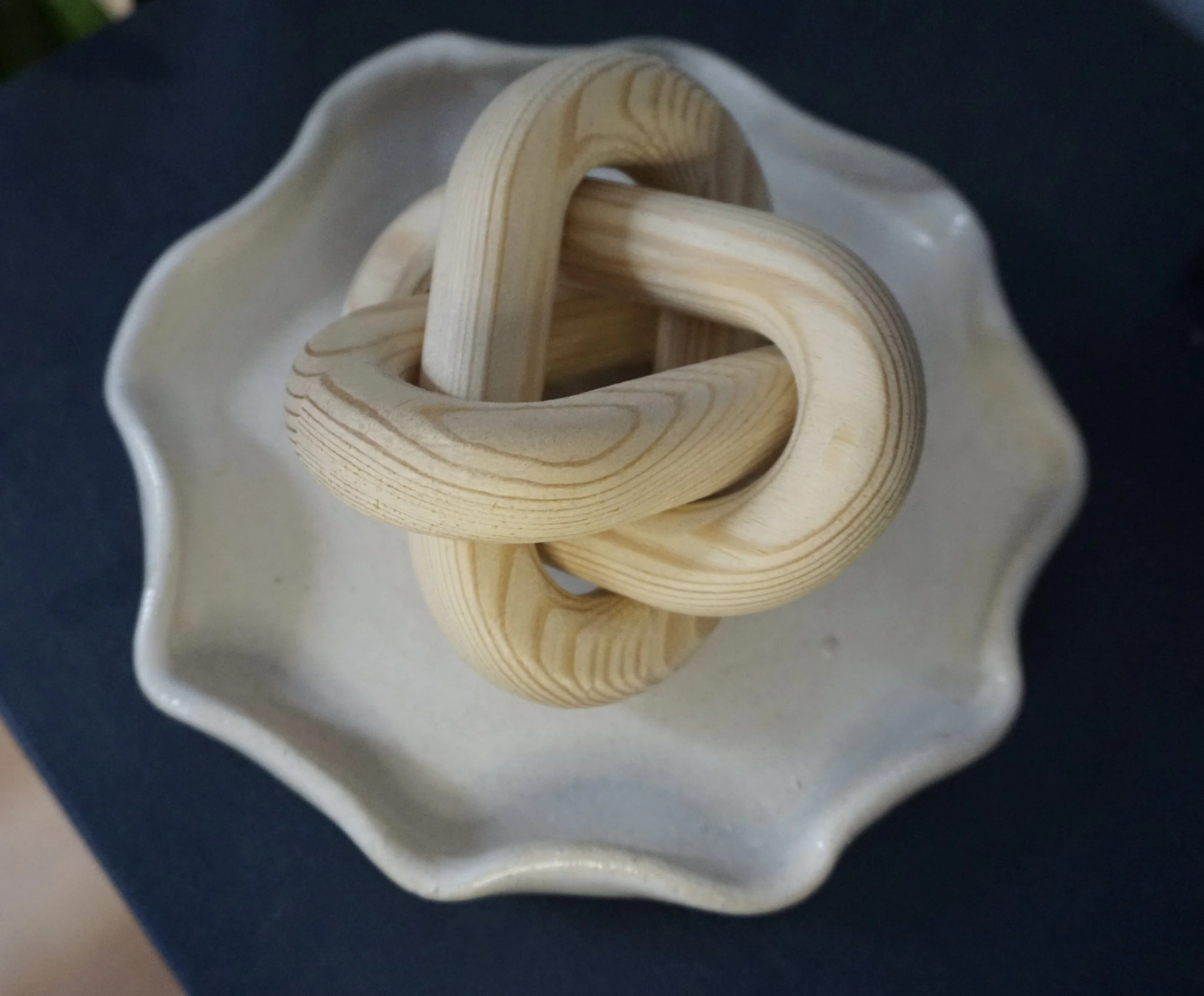 A wooden knot sculpture placed on a white ceramic dish with scalloped edges, on a dark surface.