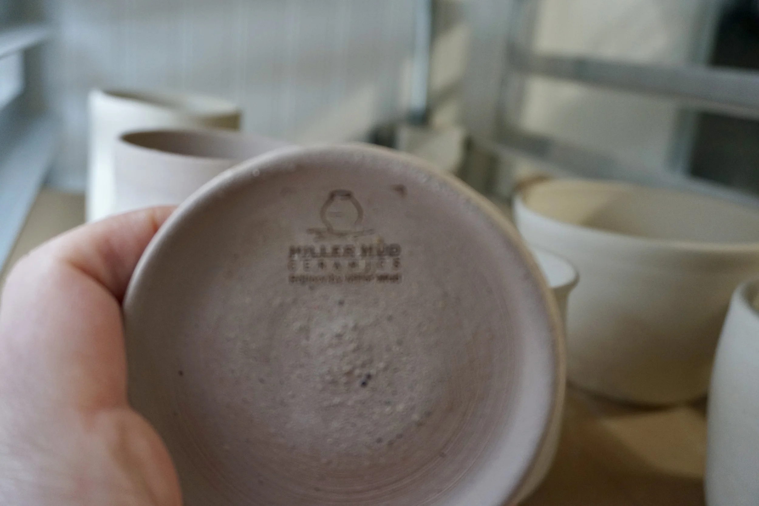 The bottom of a ceramic mug with a stamp reading 'Fuller Mug Ceramics' and a logo, held in a person's hand with other mugs in the background.