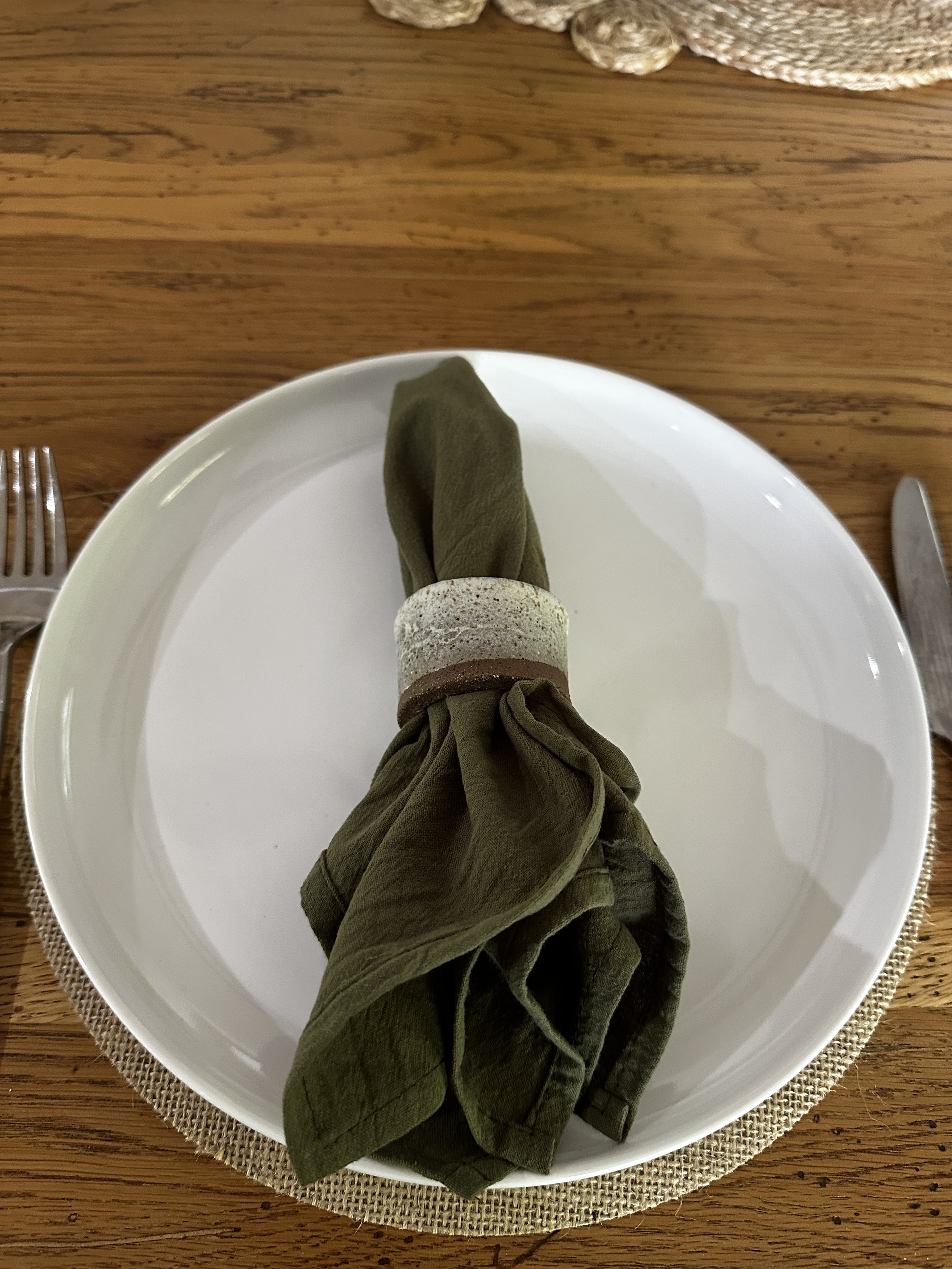 A white plate with a folded green napkin held by a ceramic napkin ring, placed on a woven placemat on a wooden table.