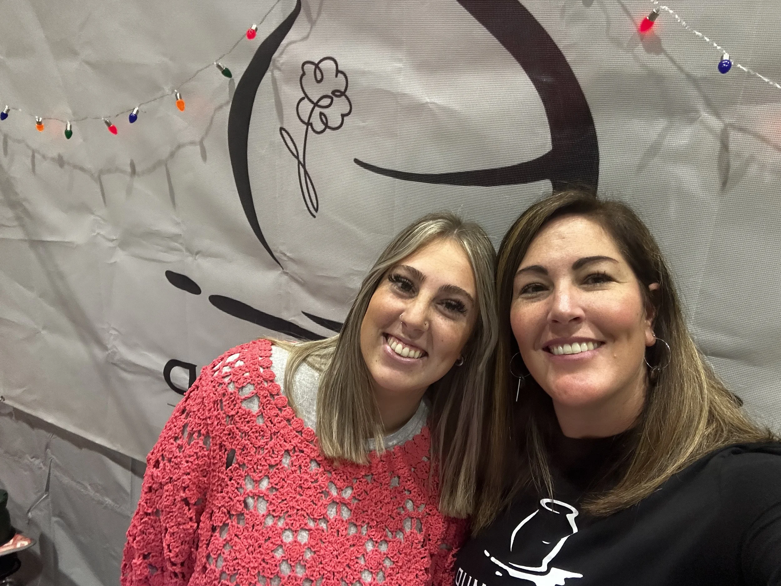 Two women smiling and taking a selfie in front of a decorated backdrop with string lights and a coffee cup drawing.