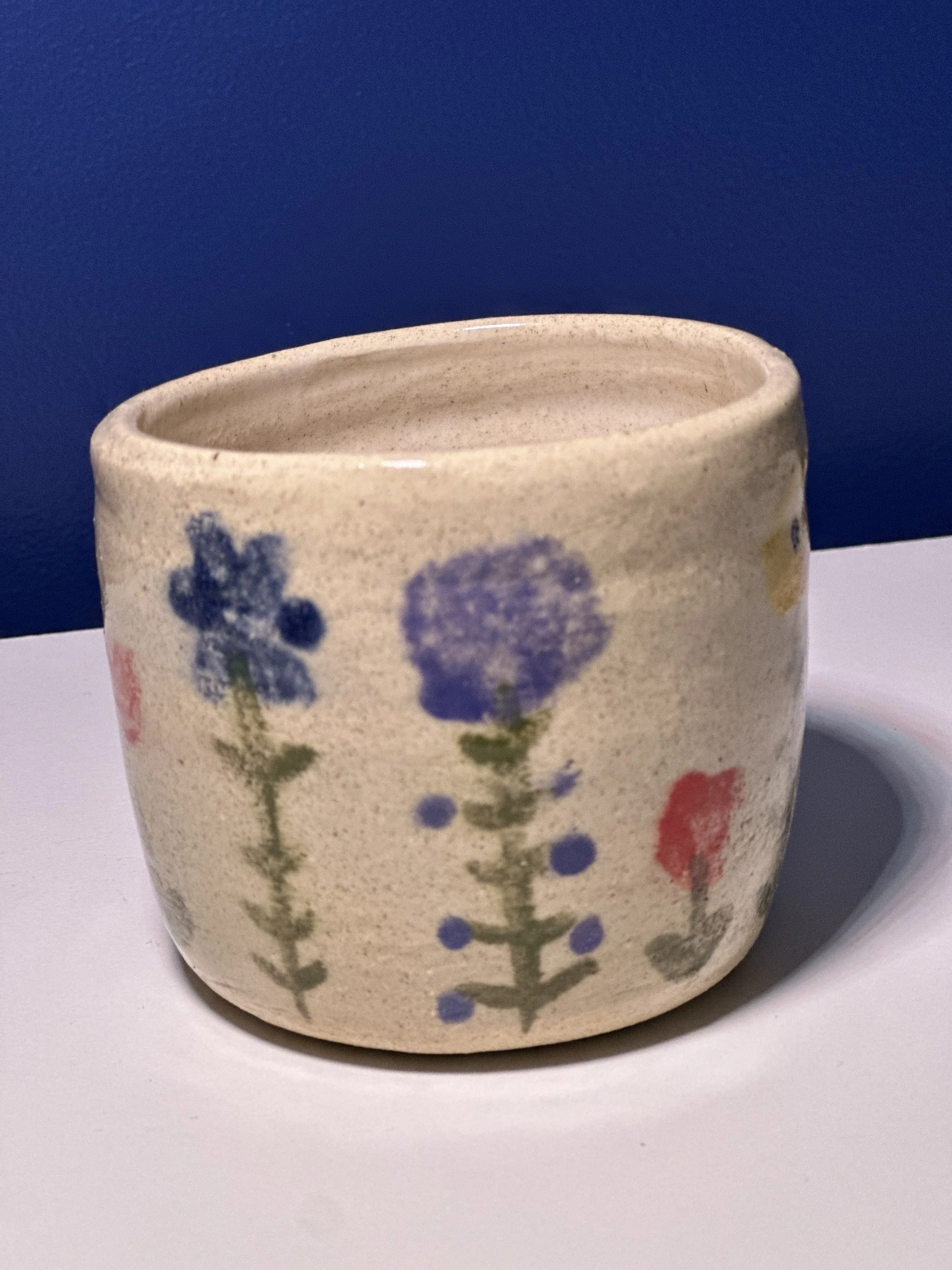 A small ceramic pot with hand-painted purple, red, and pink flowers on a beige, speckled background, placed on a white surface against a blue wall.