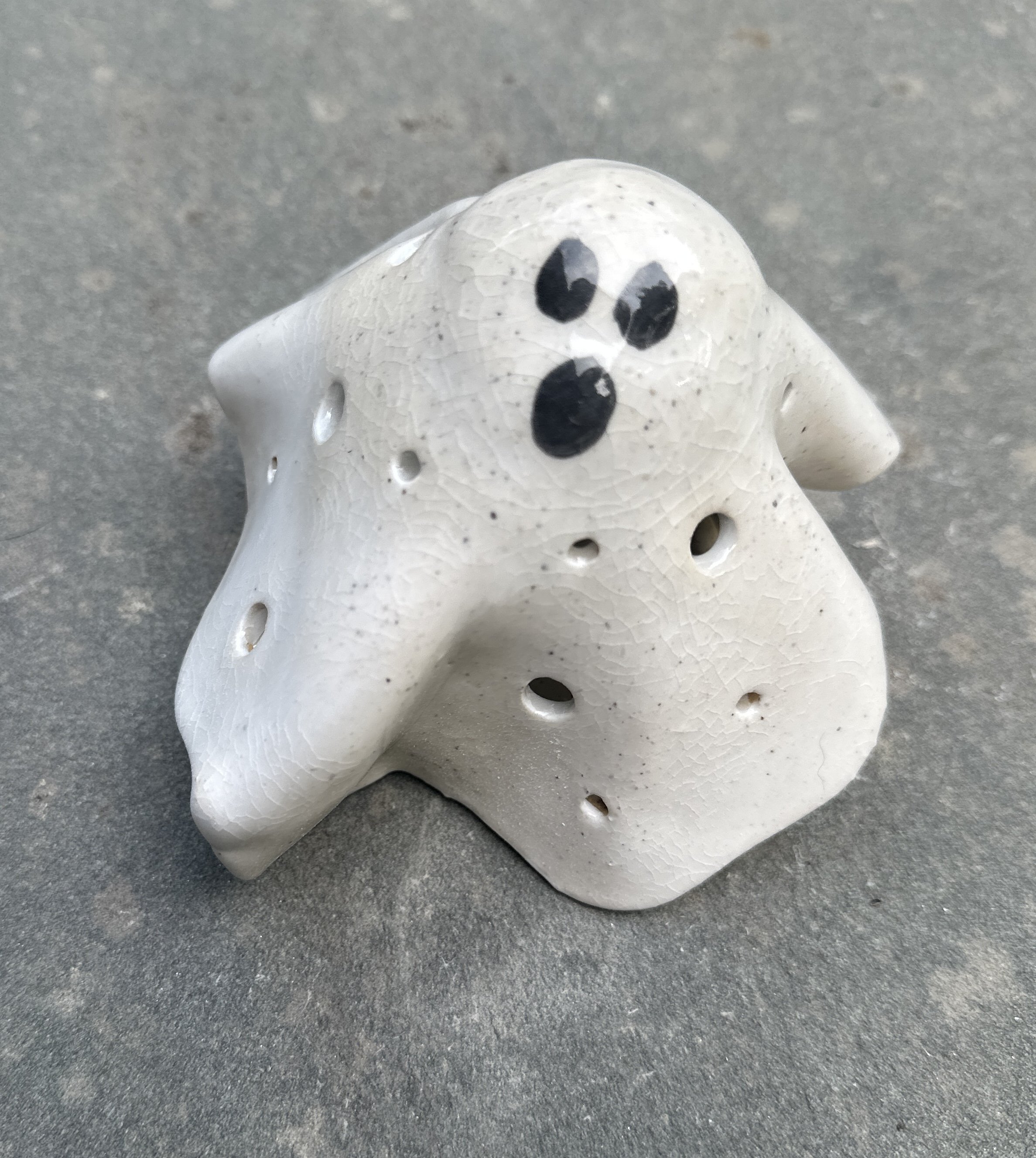 A small ceramic ghost figurine painted white with black eyes and mouth, sitting on a gray surface. The ceramic has a crackled glaze finish and holes throughout its body.