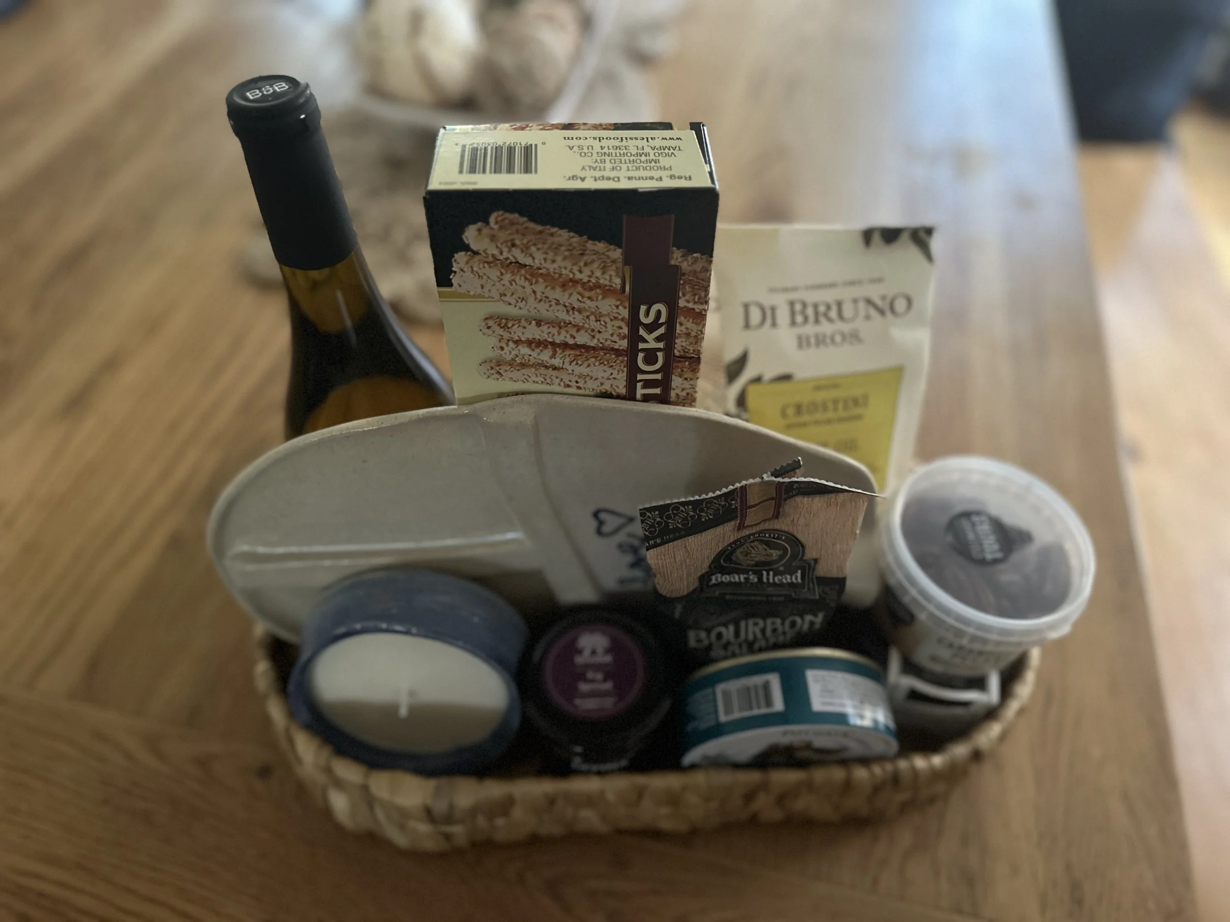A small woven basket on a wooden table containing various food items and condiments, including a bottle of mayonnaise, a box of crackers, a bag of dip mix, a container of dip, and candles.