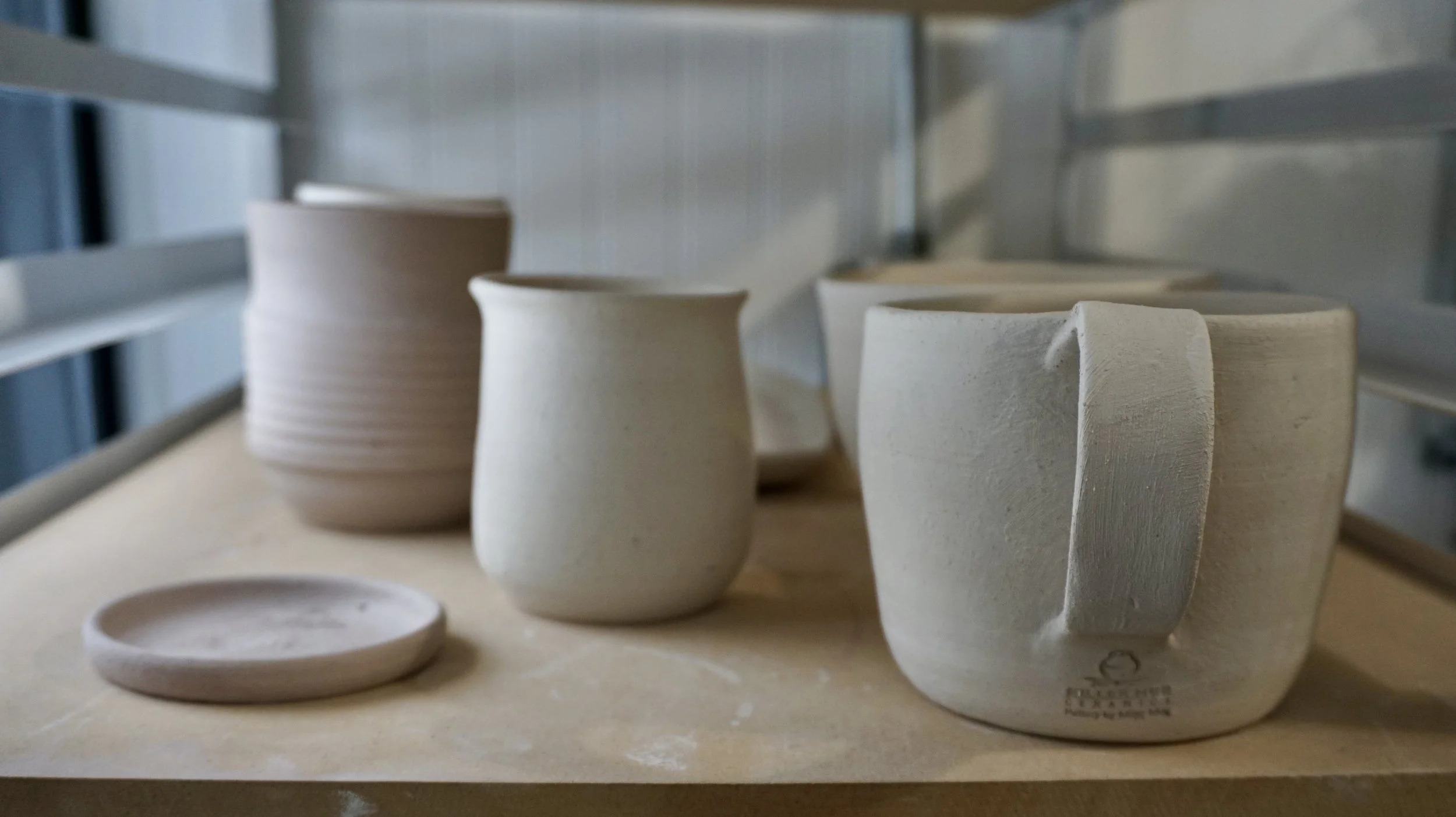 Unfinished ceramic cups and a small plate on a wooden surface, with a metal rack or shelving unit in the background.