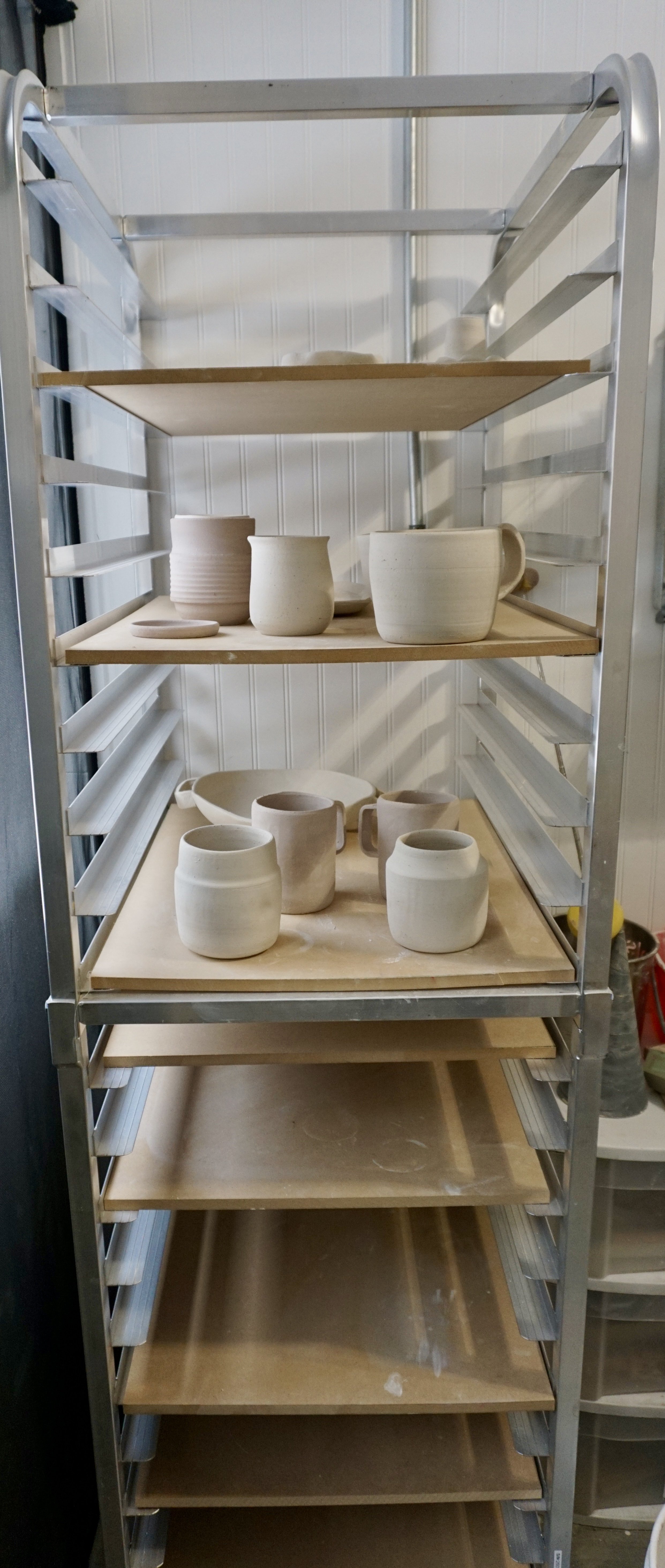 Metal shelving unit with multiple unfinished ceramic pottery pieces and plates in a pottery studio.