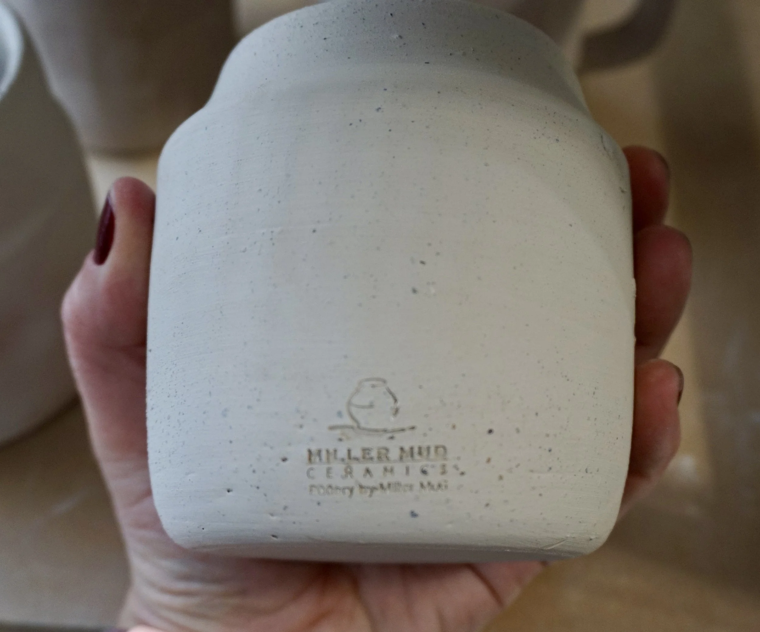 A person holding a white, textured ceramic jar with a small logo and text reading 'Hiller Mud Ceramics' on the front.