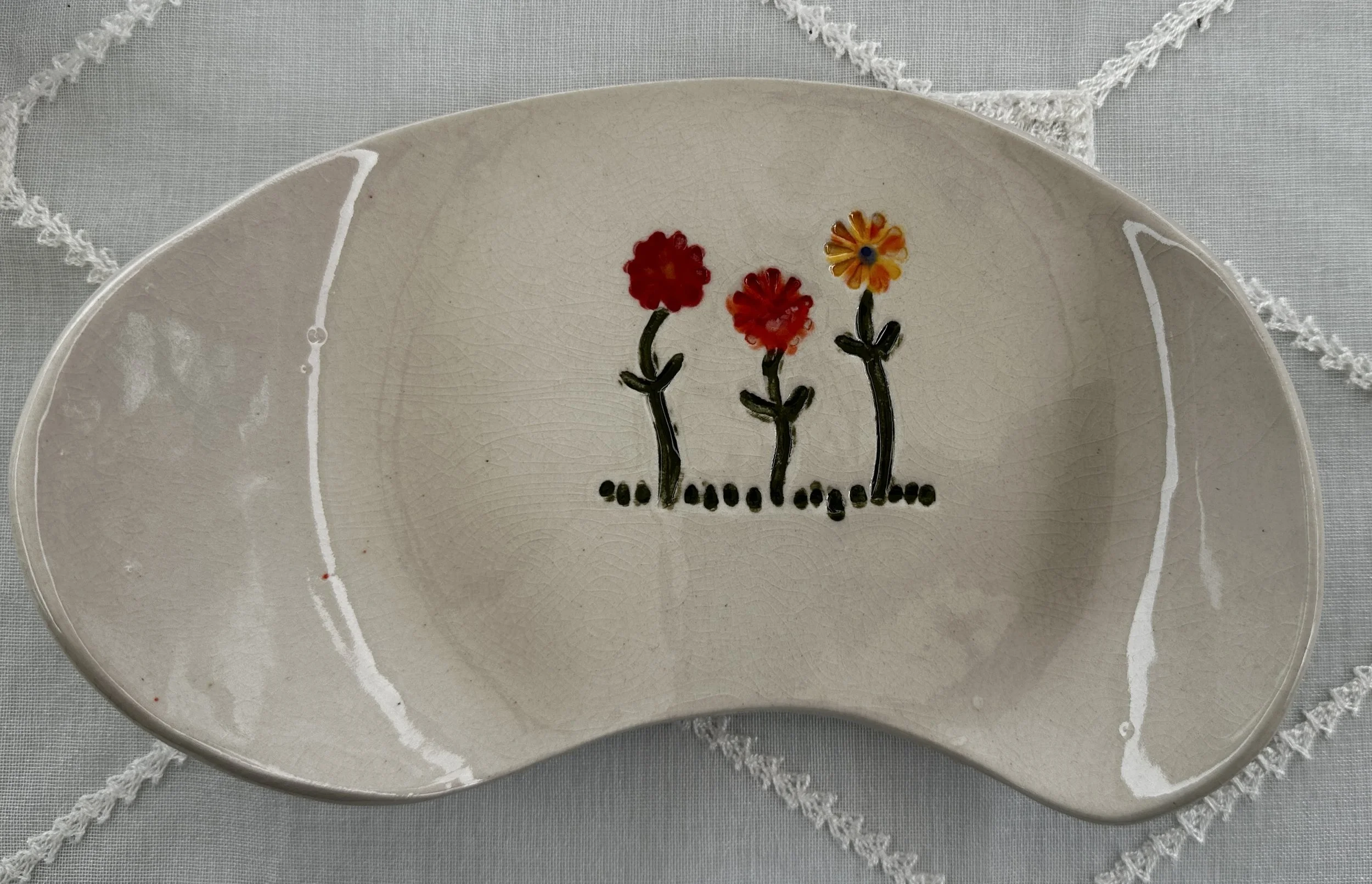 A ceramic plate with a painting of three colorful flowers and black stems, placed on a white lace-trimmed tablecloth.