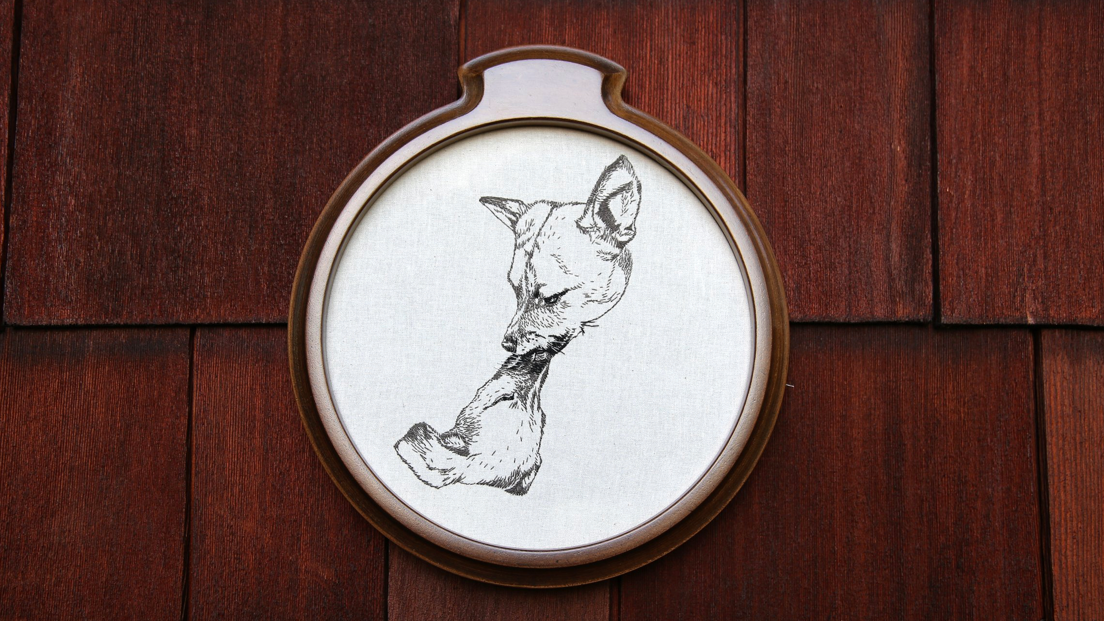 Embroidery hoop with an outline drawing of a cat's head and a curled tail on white fabric, mounted on a wooden wall.