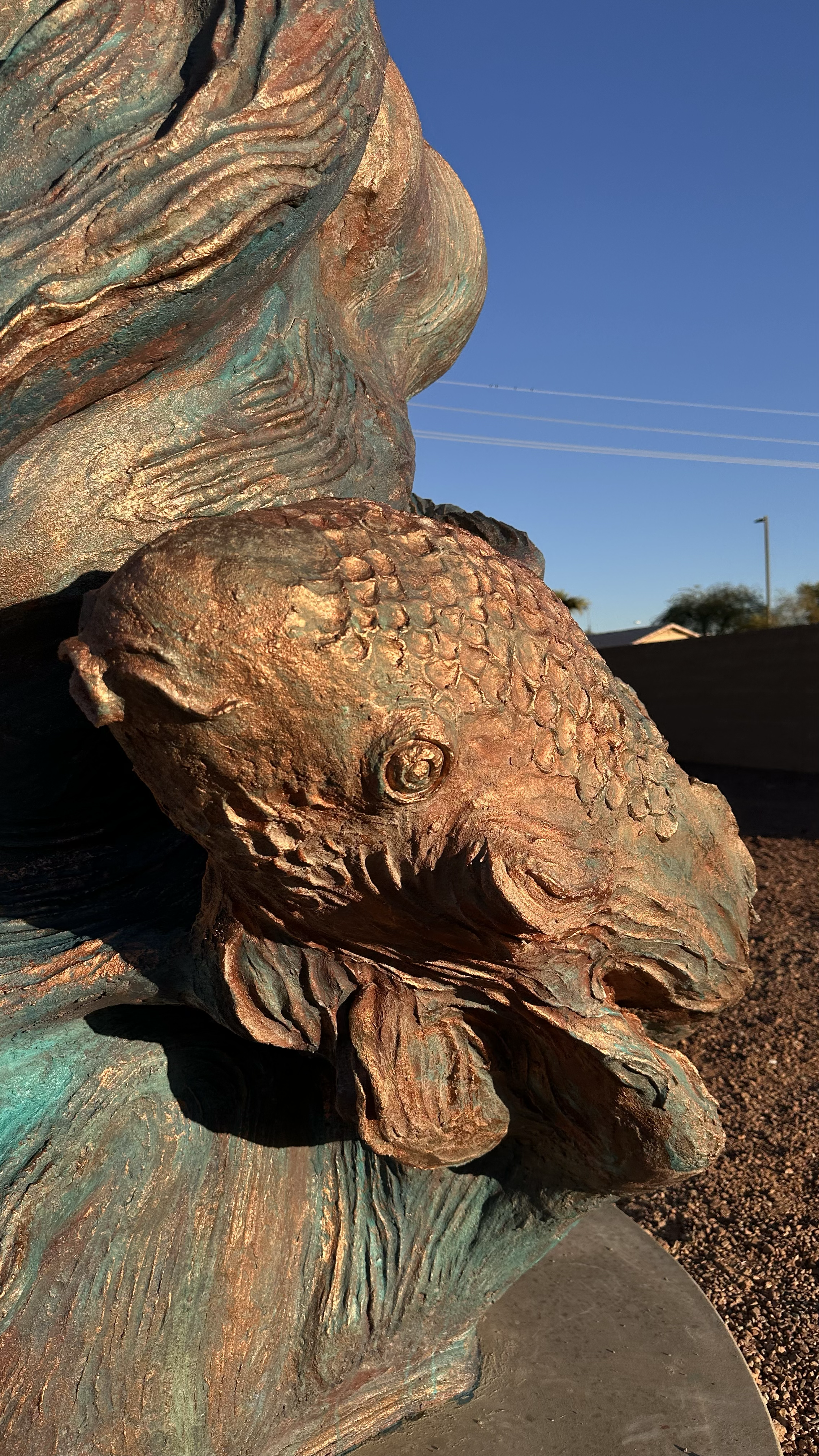 Close-up of a bronze sculpture of a fish's head with intricate details, set outdoors against a clear blue sky.