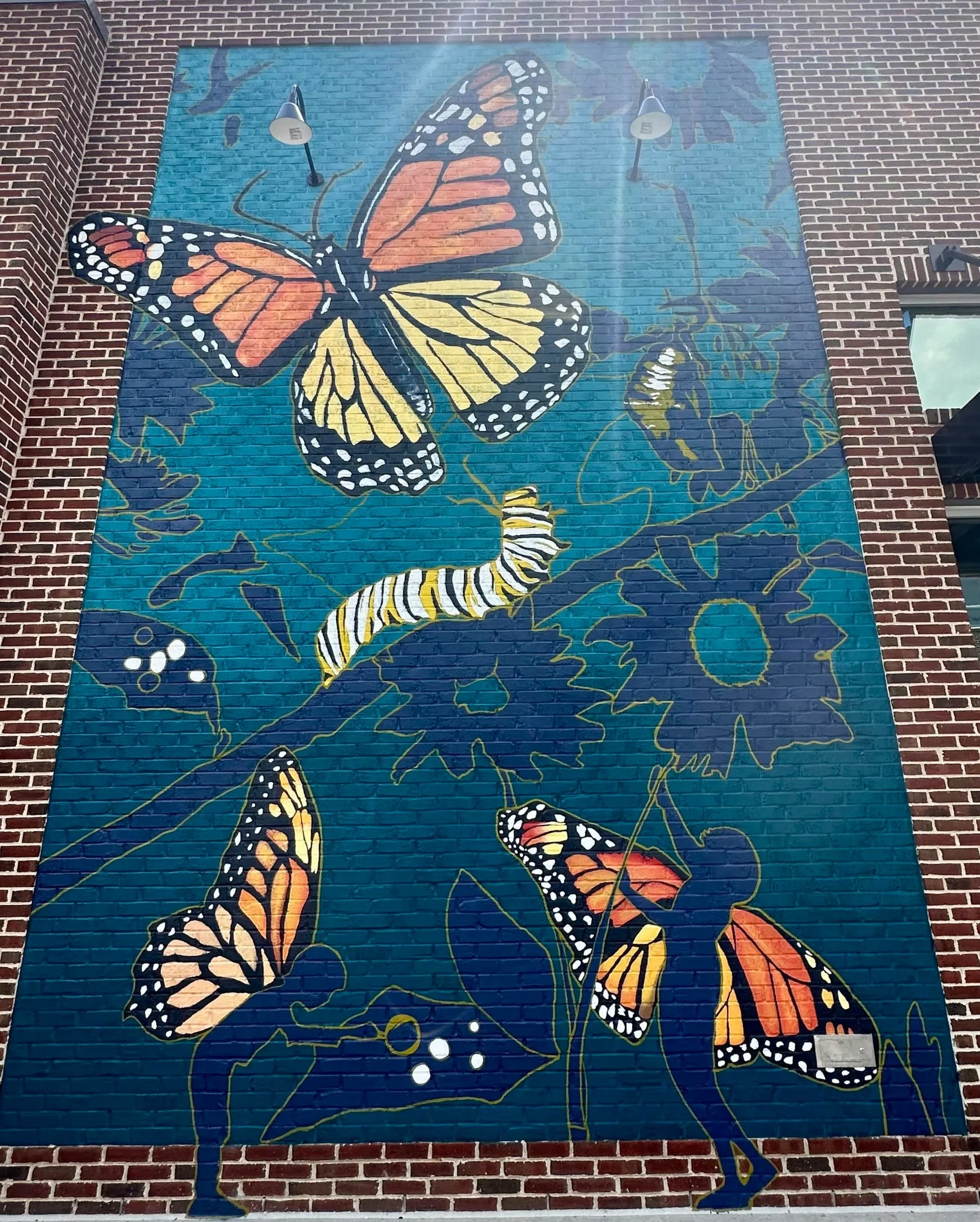 Colorful mural on brick wall featuring large monarch butterflies, caterpillars, and abstract floral designs in shades of orange, yellow, black, and white on a blue background.