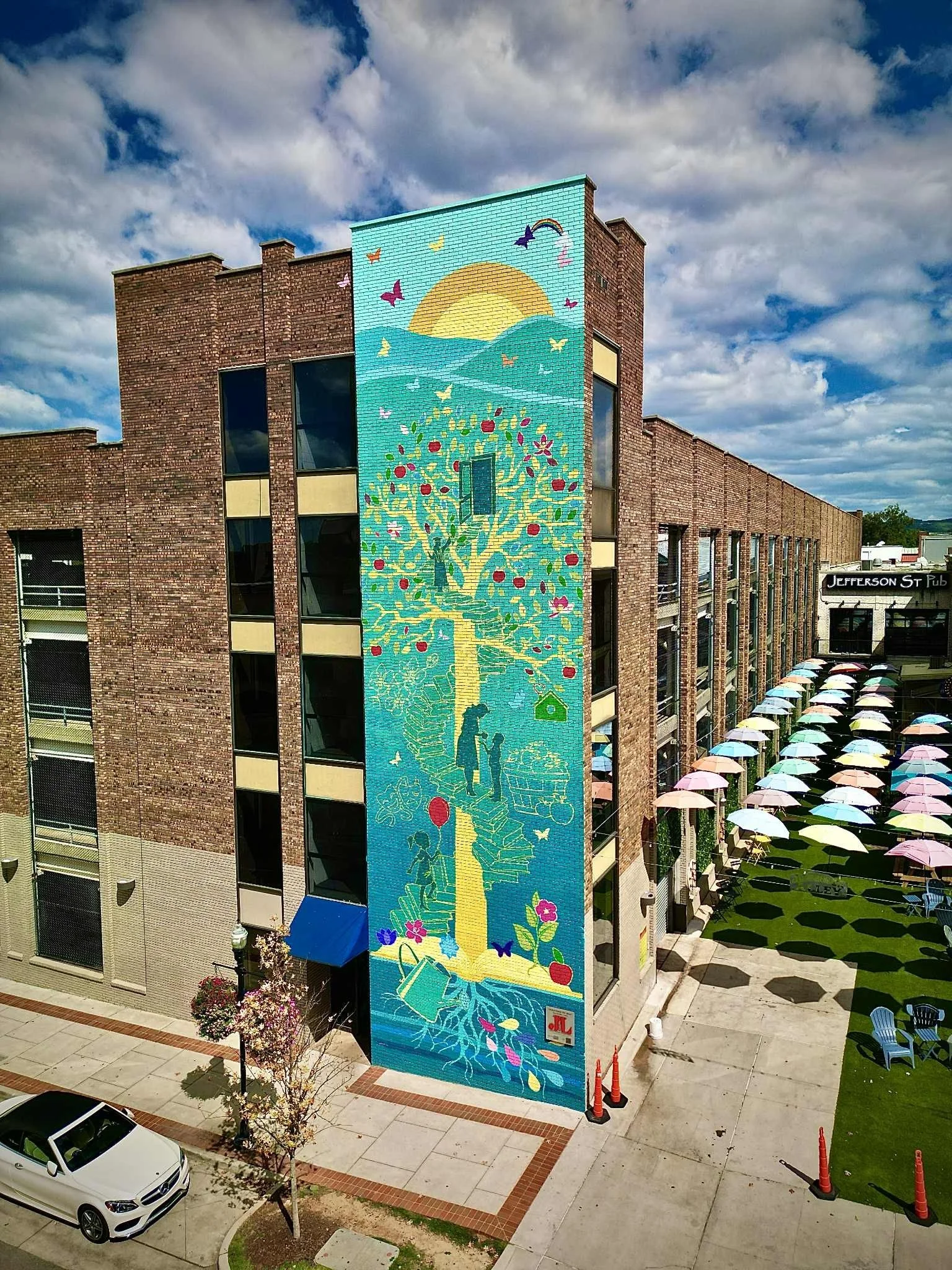 Colorful mural painted on the side of a brick building, depicting children, flowers, a tree with red apples, a sun, butterflies, and a rainbow, with a cloudy sky above.