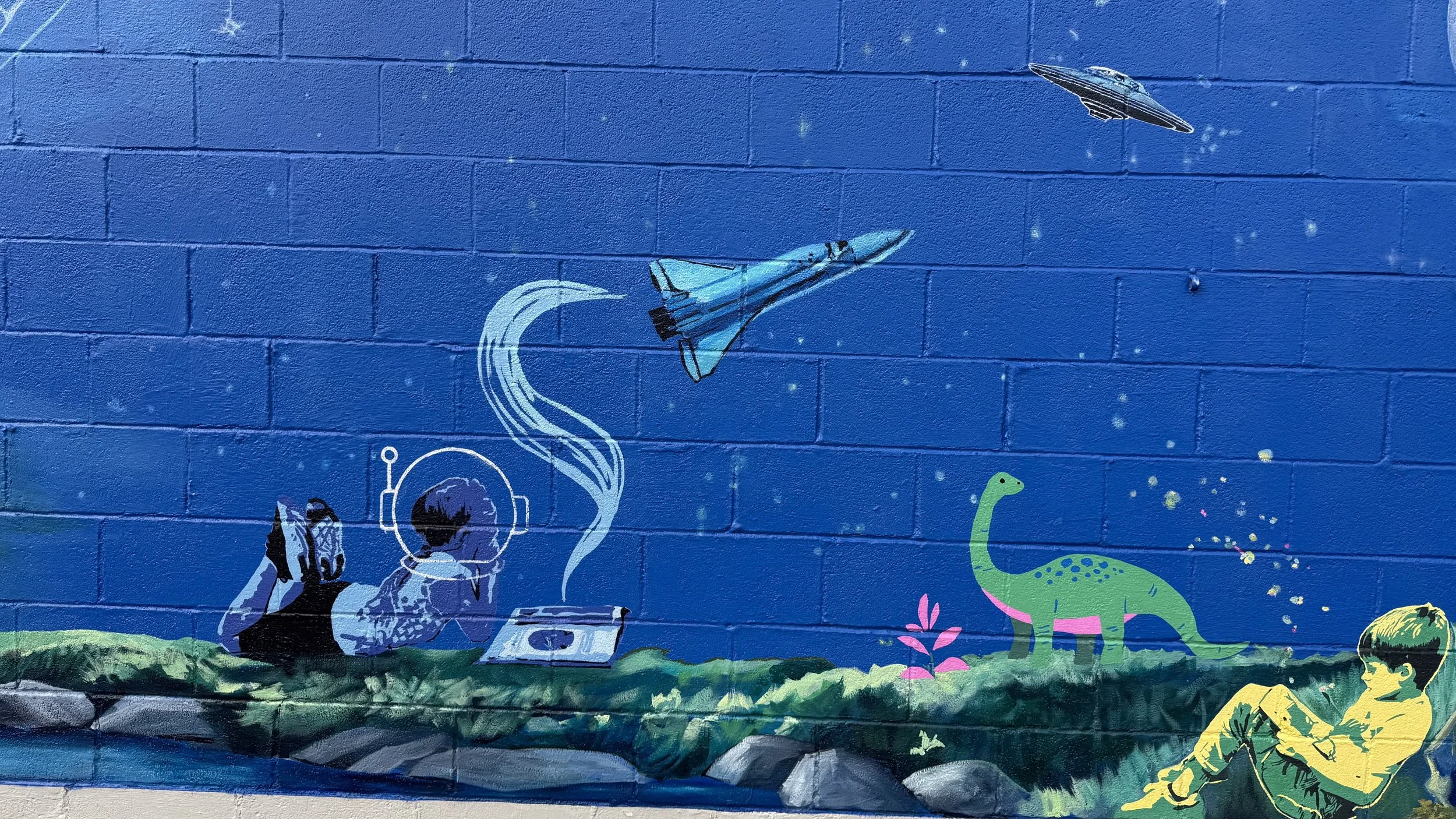 Colorful mural of a child in yellow sitting on the ground, a green dinosaur, a person with headphones, a spaceship, a rocket, and stars on a blue brick wall.
