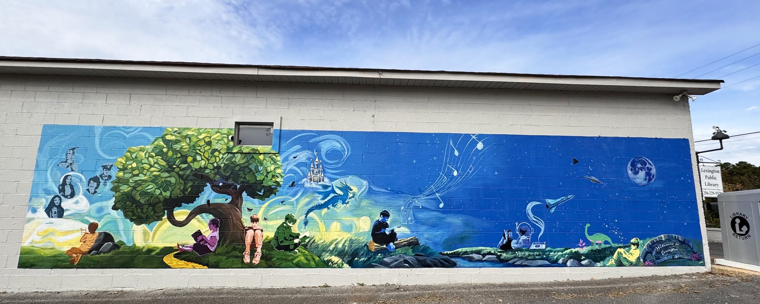 Colorful mural on a white brick wall depicting children reading books under a large green tree, with musical notes, a dragon, and a moon in a blue sky, representing creativity and learning.