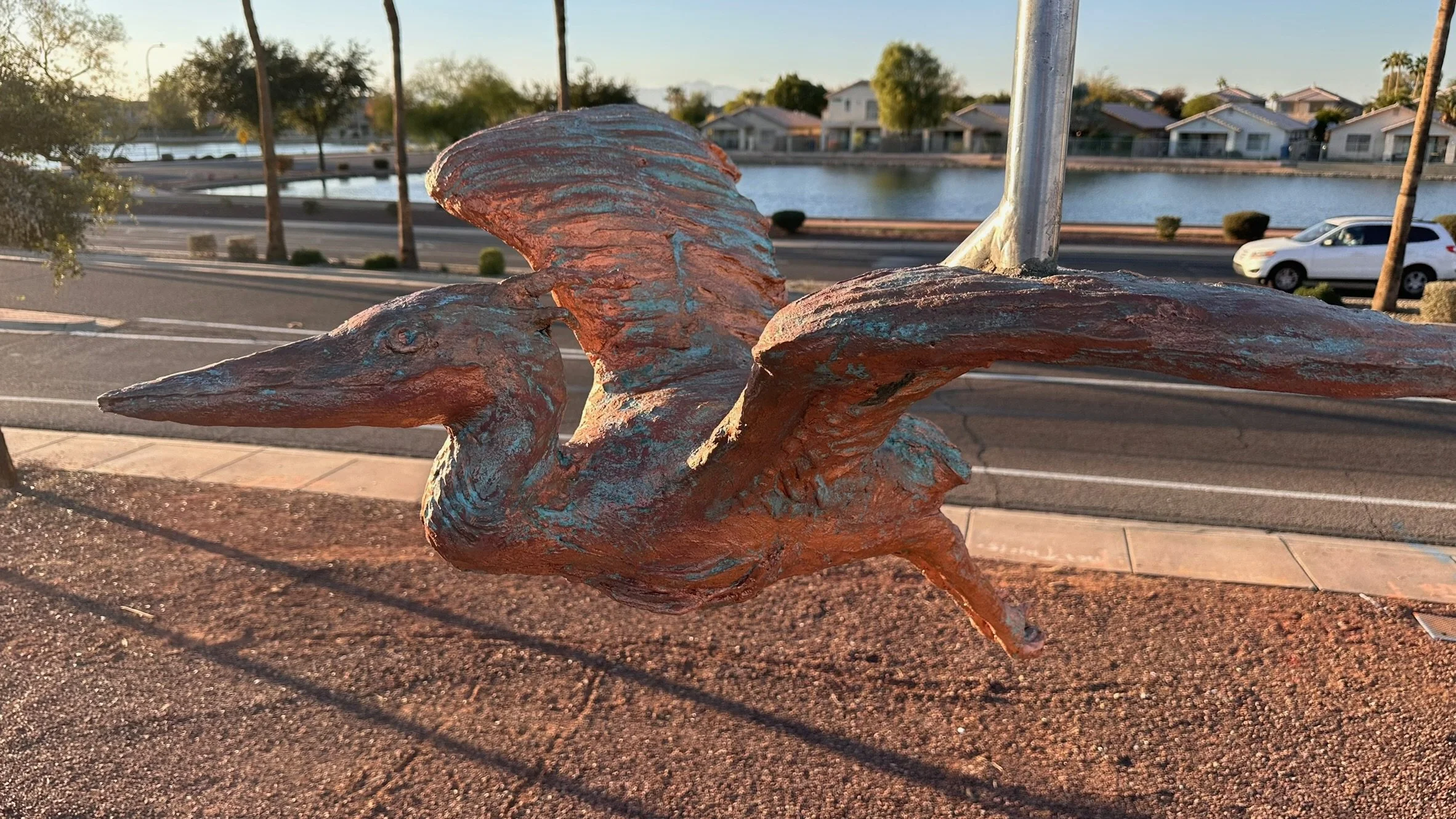 Close-up of a rusted bronze sculpture of a heron with wings extended, set outdoors near a sidewalk with a street and houses in the background.