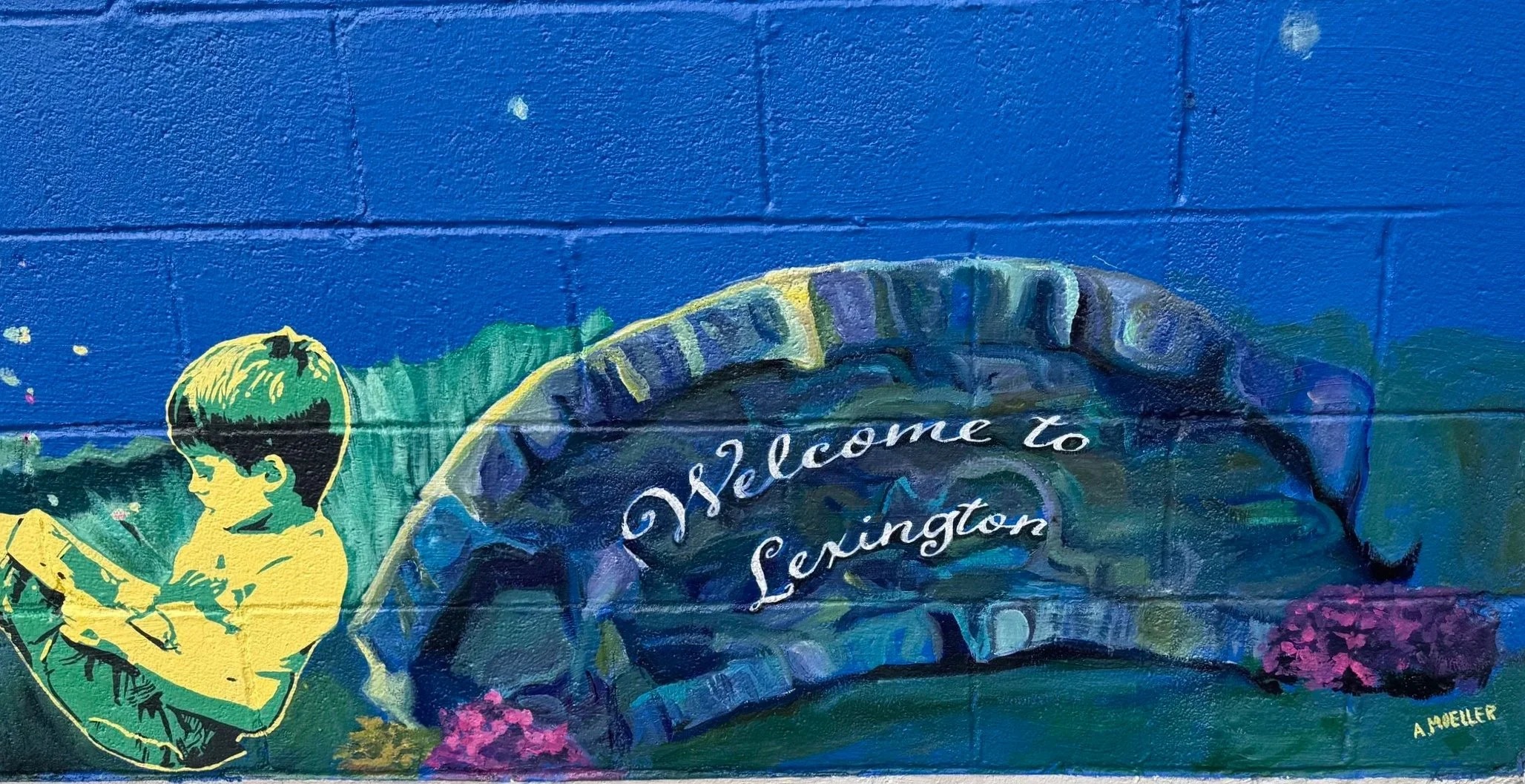 A colorful mural on a brick wall featuring a boy with glasses reading a book, a stone arch with the words "Welcome to Lexington" painted on it, set against a bright blue background with a small pink bush in front.
