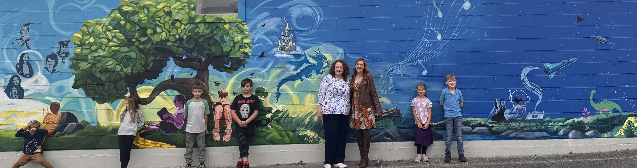 A group of children and two women standing in front of a colorful mural painted on a brick wall. The mural depicts a large tree, a castle, a dragon, musical notes, and space-themed elements like spaceships and planets.