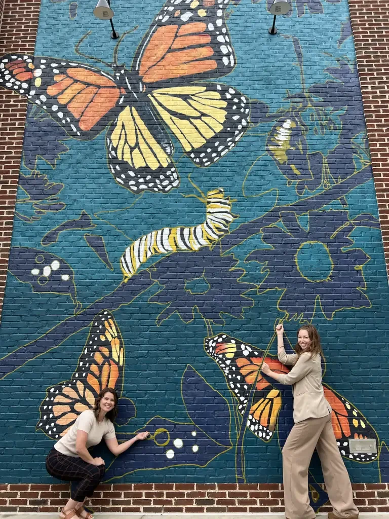 Two women standing in front of a large mural on a brick wall depicting monarch butterflies, a caterpillar, and flowers with vibrant colors and detailed designs.