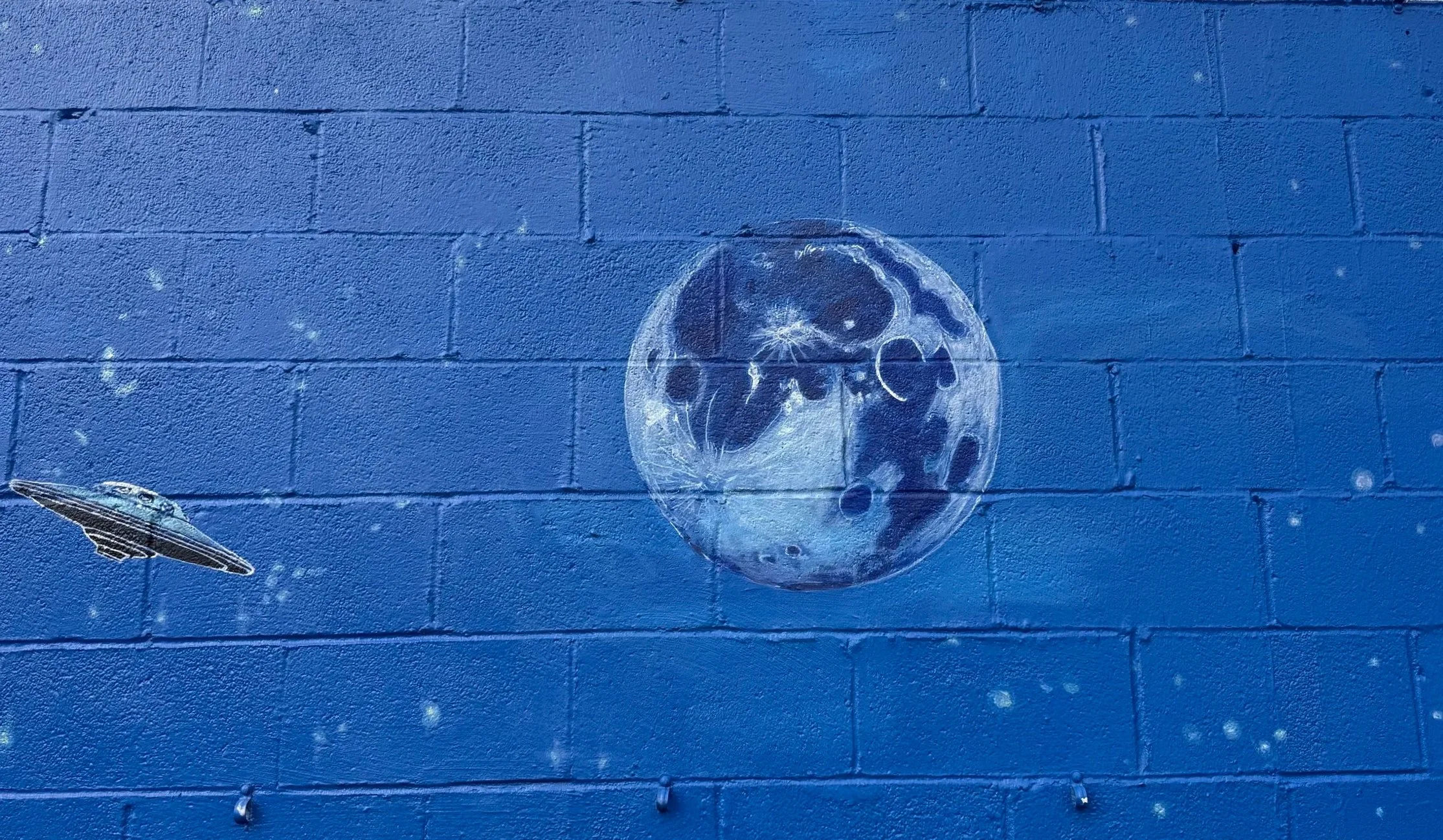 Street art mural featuring a space scene with a flying saucer, a large moon with craters, and stars painted on a blue brick wall.