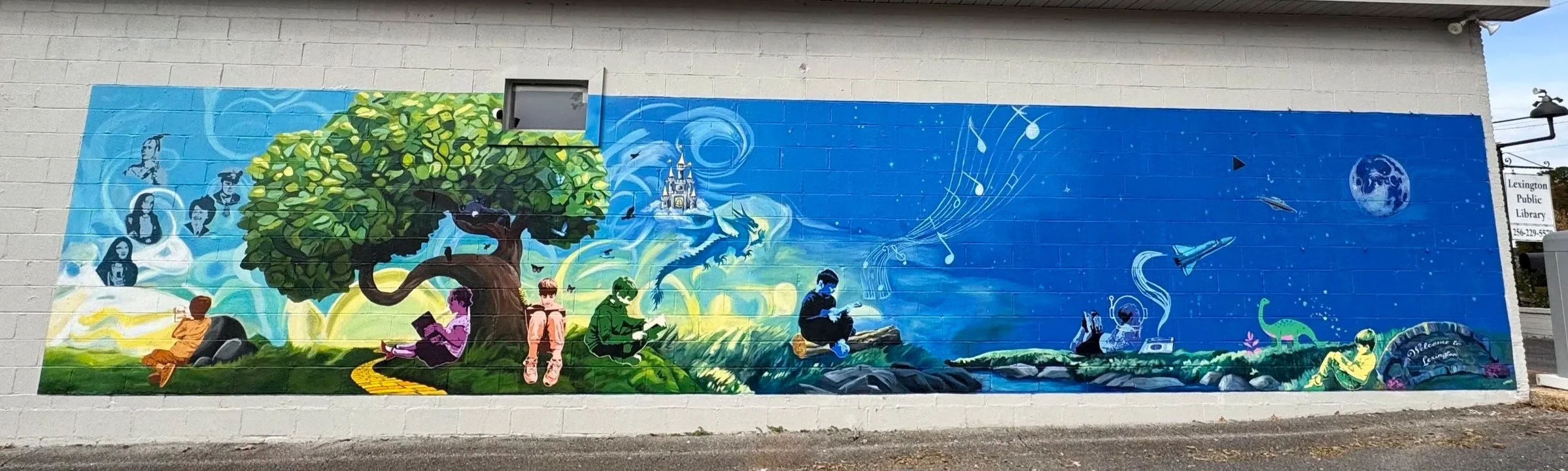 A colorful mural on a brick wall depicting children reading and playing outdoors under a large tree, with a castle in the background and scenic elements like a dragon, a spaceship, musical notes, and planets.